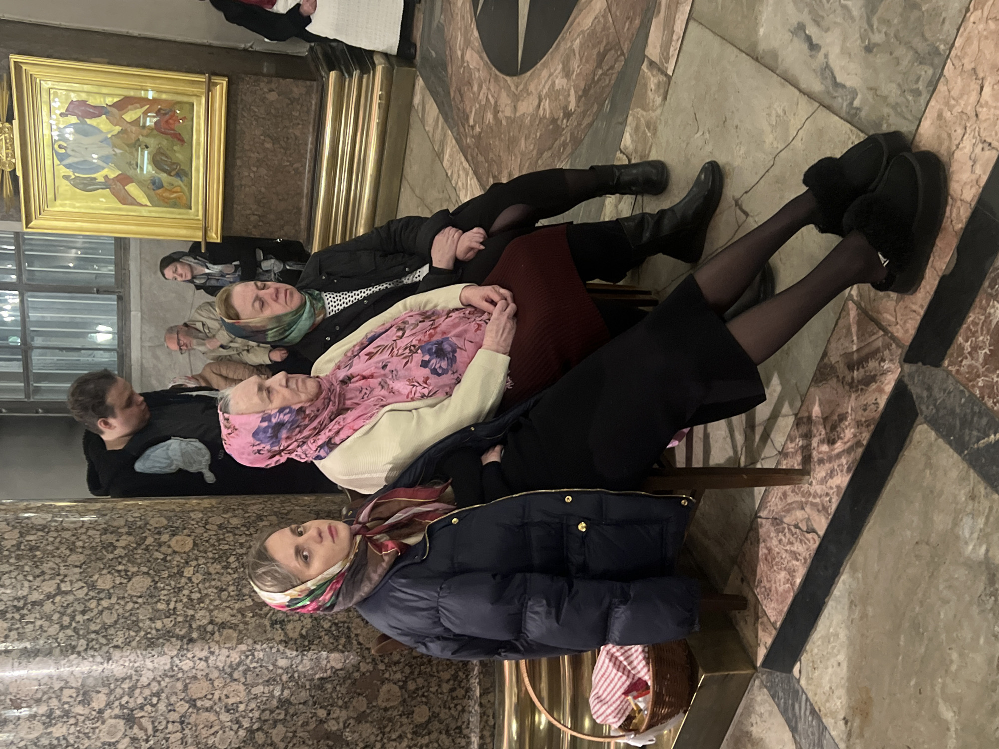 The image shows a group of women sitting in the Church of the Savior on Spilled Blood in Saint Petersburg, Russia. Three women are prominently visible in the foreground, each wearing headscarves. The woman closest to the viewer is wearing a dark blue puffer jacket and a patterned scarf. Behind her, another woman is wearing a light beige cardigan and a pink floral headscarf. Further behind, a third woman is wearing a black jacket and a patterned headscarf. Other people are visible in the background, including men and women. A painting in a gold frame hangs on the wall. The floor is made of marble with a geometric pattern.