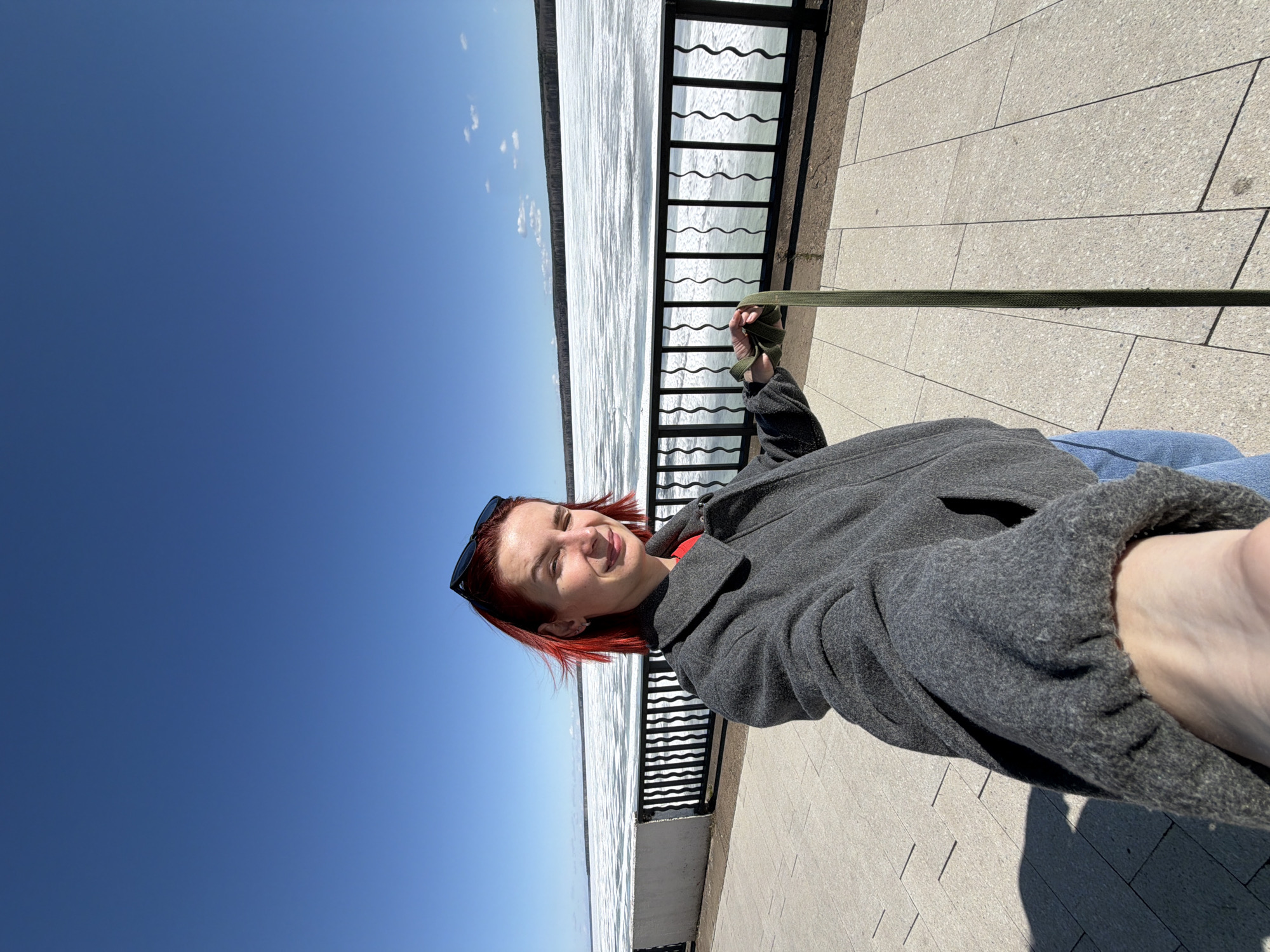 A woman with red hair and sunglasses stands outside on a sunny day in Chaikovsky, Russia. She is holding a green strap or line with her right hand, and is wearing a gray coat and blue jeans. A black metal railing separates her from the view, which includes a body of water that appears to be frozen or partially frozen, and a blue sky with a few clouds. The ground is paved with gray rectangular stones.