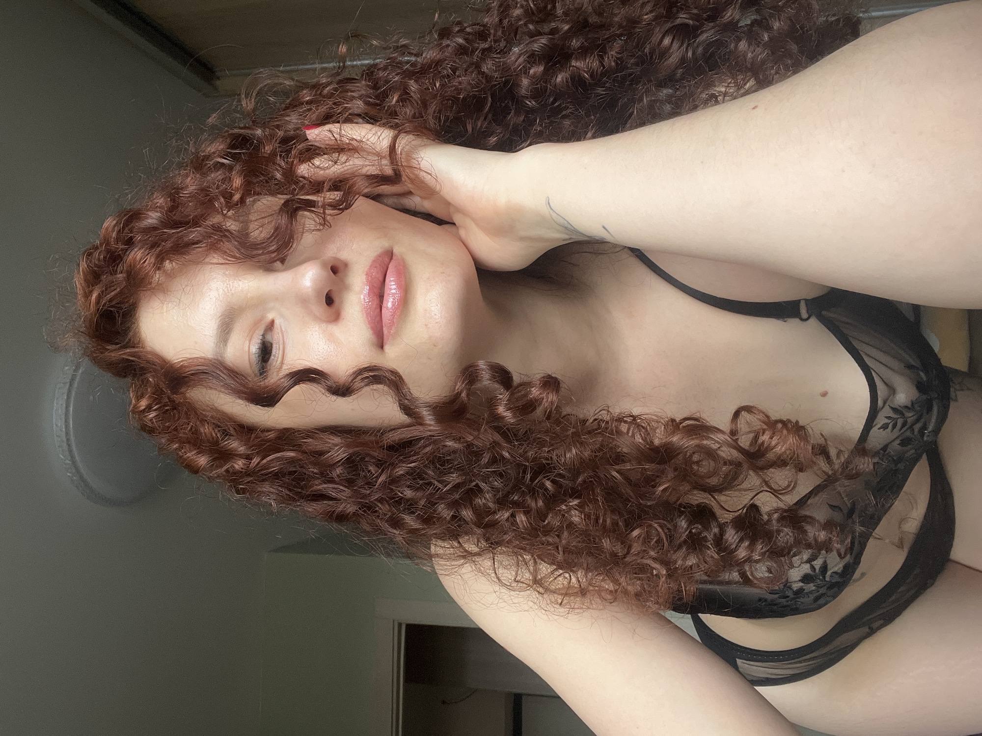 A person with curly red hair is shown in a selfie wearing black lingerie. The background features a gray wall, a round object, and a doorway. The exact location within Saint Petersburg, Russia, cannot be determined from the image alone.