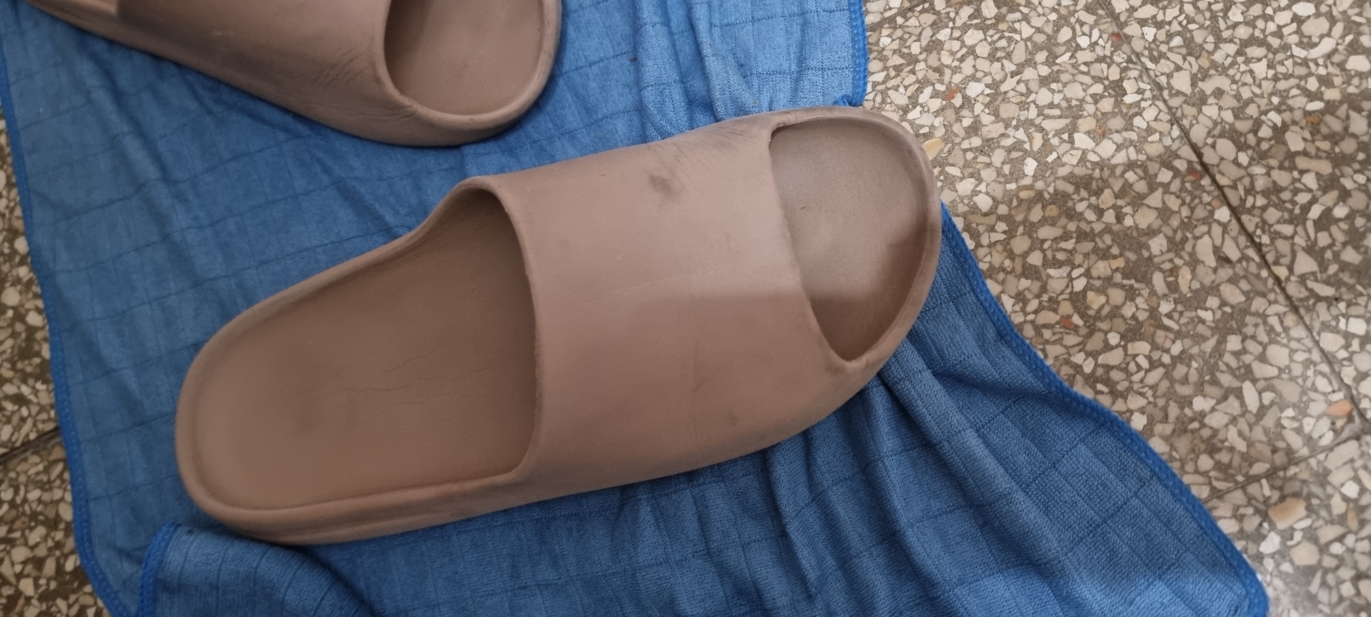 A close-up, high-angle shot shows a pair of light brown slides lying on a textured blue microfiber cloth. The slides are positioned diagonally, with the left slide more prominently featured, angled towards the viewer's lower right. The surface of the slides appears somewhat worn, with subtle lines and a few faint smudges. The blue cloth has a grid-like texture and is laid out on a mottled, terrazzo-like tiled floor, which is visible in the background and in the lower right corner of the frame. The lighting suggests an indoor setting, possibly a home or a shoe store. The city of Kiryat Bialik, Israel, is a residential area known for its parks and community centers; this scene is consistent with a domestic environment within such a city.
