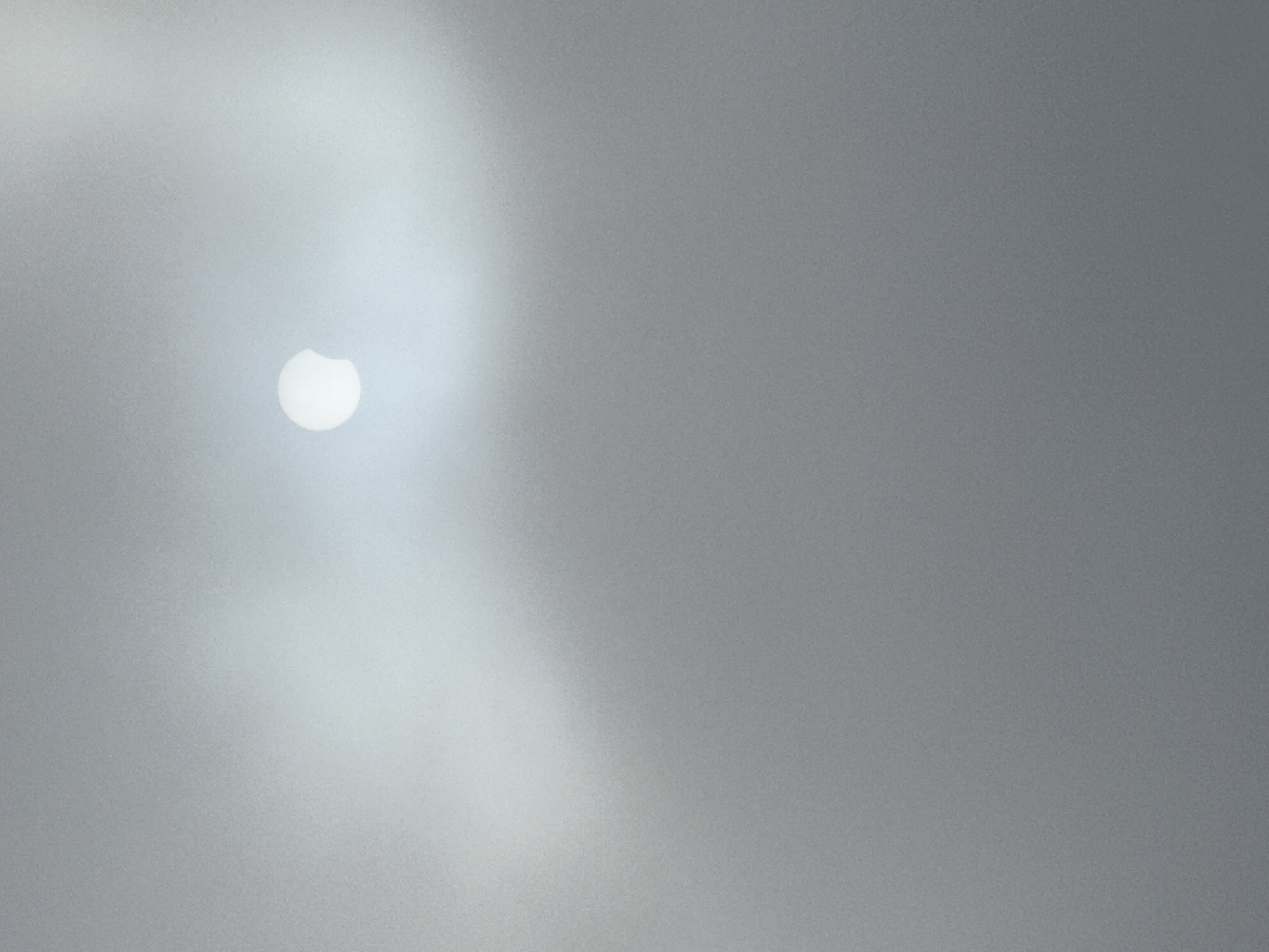 The image shows a partial solar eclipse viewed through a hazy or cloudy sky. A bright disc, partially obscured by a darker circular shape, is visible in the upper portion of the frame. The rest of the image is filled with a diffuse, gray haze, suggesting overcast conditions. There are no people, objects, or specific landmarks identifiable within the frame, making it impossible to determine the location in Vila Nova de Gaia, Portugal, or any other specific city. The scene is abstract and lacks any contextual details.
