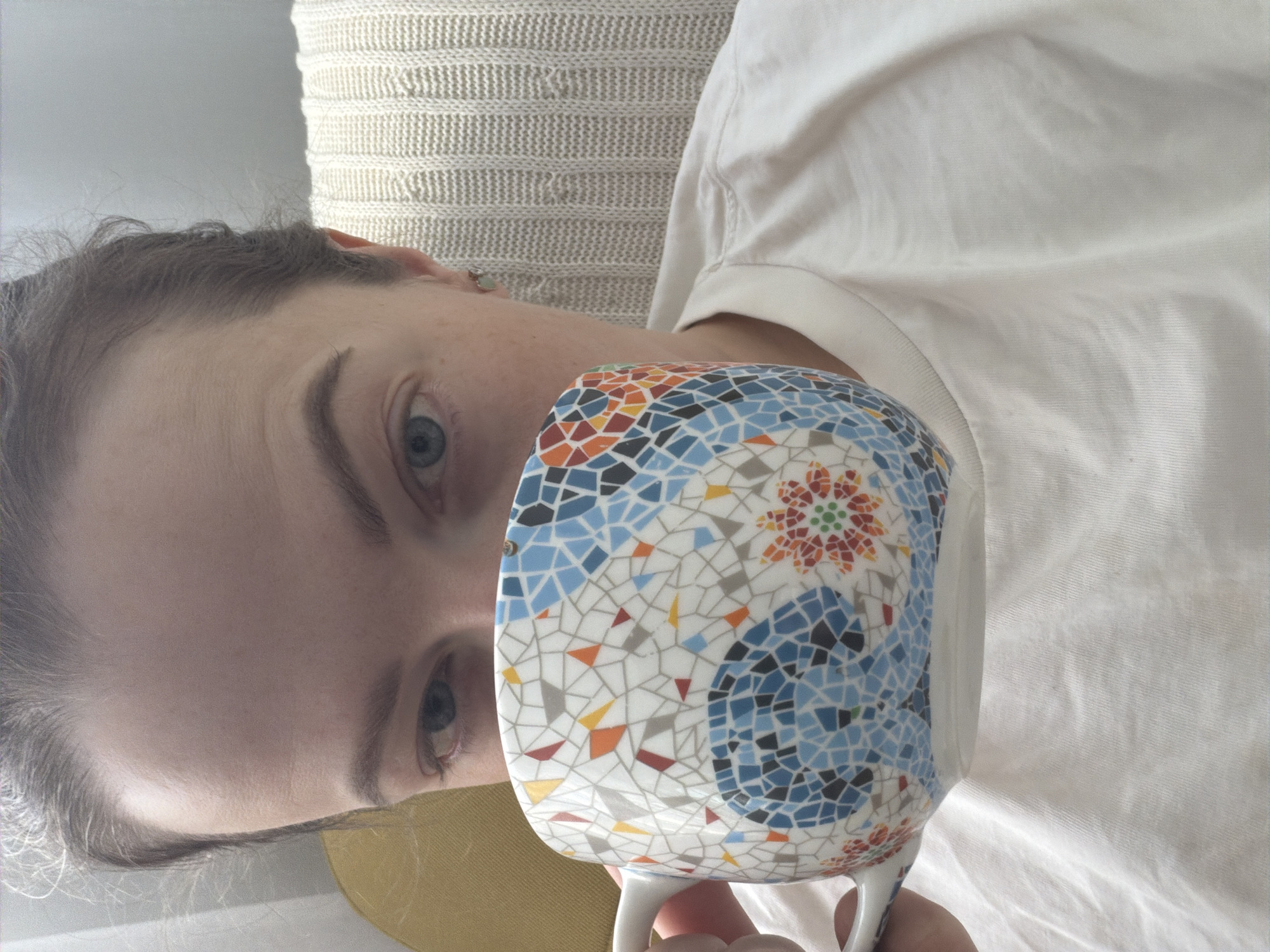A close-up, portrait-style photograph depicts a person, likely female, with fair skin, blue eyes, and subtle freckles, looking directly at the viewer. Her dark, wavy hair is visible around her forehead. She is wearing a light-colored, possibly white or cream, top.
A distinctive white mug with a vibrant mosaic pattern is held in front of her mouth and lower face, partially obscuring it. The mosaic design on the mug consists of irregularly shaped tiles in various shades of blue, orange, red, black, grey, and white, with a notable red and orange floral or star-like motif on its side.
The background includes a light-colored, textured knitted fabric, possibly a cushion or blanket, behind her head and shoulder. Another plain light-colored fabric is visible to her right, and a hint of a yellow or mustard-colored fabric is seen near her neck.
The person appears to be in a relaxed, casual moment, either about to drink from or having just taken a sip from the mug. The lighting is soft and natural, indicating an indoor setting during the daytime. Her expression is neutral and contemplative. No text is visible in the image. The location context for this image is Moscow, Russia, though no specific cues are visible within the frame.