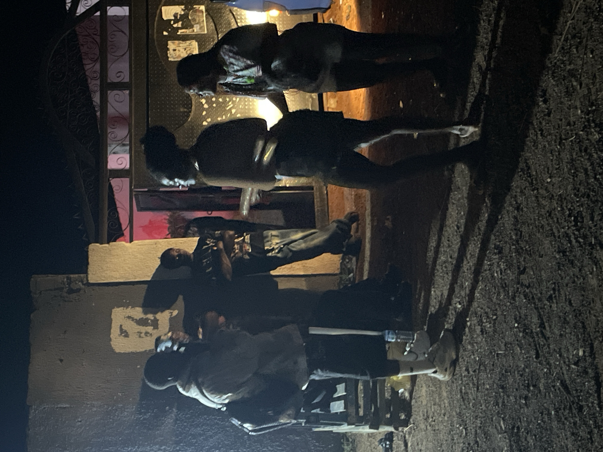 The image depicts a nighttime scene in Durumi, Nigeria, where a group of people are gathered outdoors. The setting appears to be a slightly unkempt area with dirt ground, suggestive of a residential or communal space. A building with a rough-textured wall and a gated entrance is visible in the background.
In the foreground, four individuals are the primary focus. On the left, a person wearing a light-colored hoodie and dark pants with a backpack on is facing towards the right, appearing to be in conversation with others. Next to them, another person is visible, partially obscured.
To the right, two individuals are engaged in interaction. One is a woman with her hair styled up, wearing a dark outfit, and she is looking to her left. Another woman, to her right, is wearing a patterned top and dark pants, with her arm extended as if touching the first woman. Both are facing towards the left of the frame.
Various objects are present in the scene, including a water bottle on the ground near the person in the hoodie, and a wooden structure, possibly a crate or bench, to the left. The lighting is dim, suggesting artificial light sources, possibly from a nearby building or vehicle, creating strong shadows on the ground. The overall atmosphere appears casual and informal.