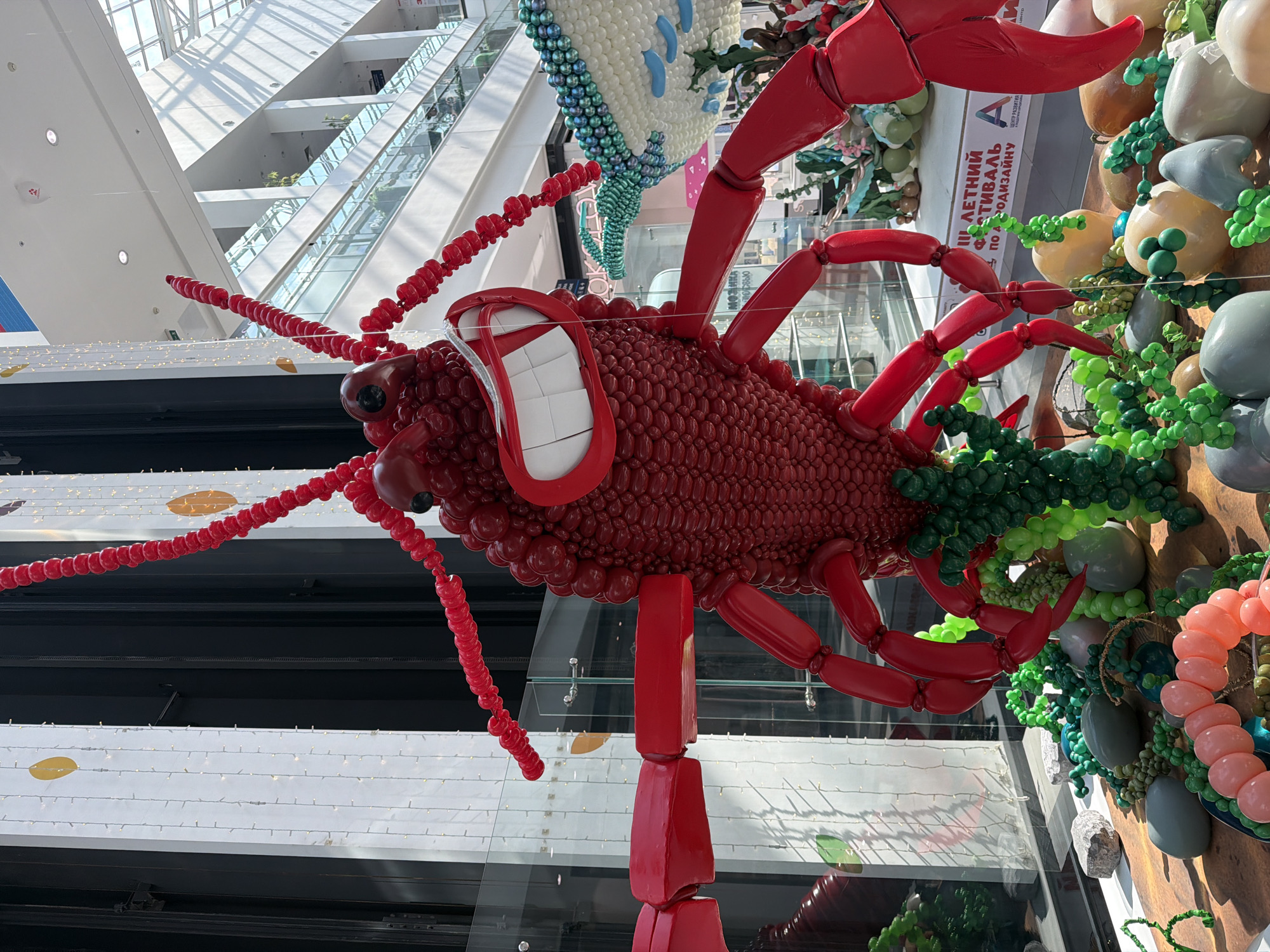 A large, vibrant red sculpture of a cartoon-like lobster, composed primarily of numerous small, round elements, dominates the foreground. The lobster features exaggerated white teeth, black eyes, long segmented antennae, and prominent claws, all in red. The sculpture appears to be made from balloons or similar inflatable materials, creating a playful and whimsical appearance.
To the right and above the lobster, other colorful balloon-based sculptures are visible, contributing to an overall marine-themed art installation. These include a white and blue jellyfish-like creature and various clusters of green, grey, yellow, and pink spherical shapes, reminiscent of coral or underwater flora.
The setting is a brightly lit, spacious, multi-level indoor environment, characteristic of a modern shopping mall or large atrium. The background features clean white architecture, multiple floors connected by escalators, and glass railings, with natural light streaming in from overhead windows or skylights, indicating it is daytime.
A partially obscured sign with Cyrillic text is visible to the right. The discernible parts read "ЛЕТНИЙ ФЕСТИВАЛЬ" (SUMMER FESTIVAL) and "ПО АЭРОДИЗАЙНУ" (BY AERODESIGN), suggesting that the installation is a balloon art display for a summer festival. The overall atmosphere is festive and visually engaging.