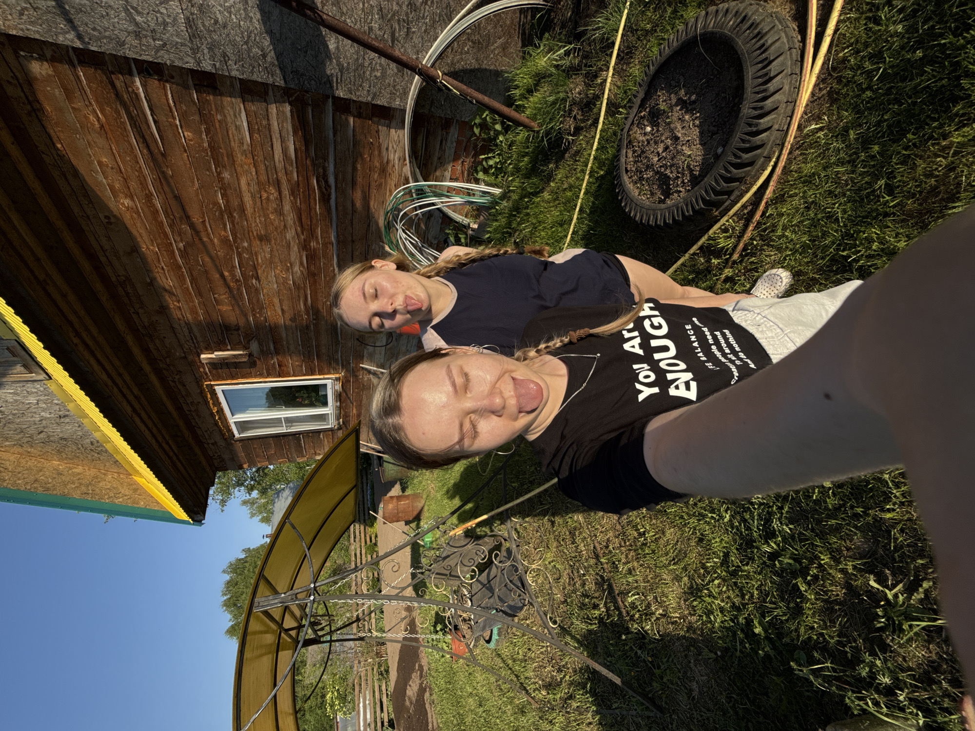 The image shows two young women posing for a selfie outdoors. They are sticking their tongues out. One woman is wearing a black t-shirt with white text that reads "You Are ENOUGH." They appear to be in a yard next to a wooden building. The yard contains a variety of items, including tires, hoses, and a metal structure that may be part of a greenhouse. The sky is visible in the background, and the lighting suggests it is daytime. The location is Zhigalki, Russia.