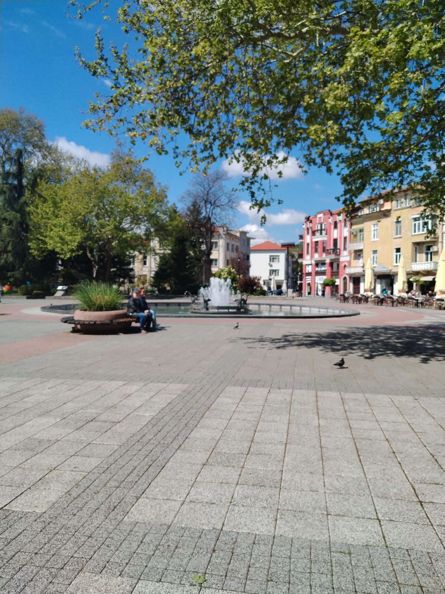This media file presents a vibrant, sunlit public square in Plovdiv, Bulgaria. The scene is captured during the day, under a clear blue sky dotted with scattered white clouds, indicating pleasant weather.
The foreground features an expansive paved area made of light and dark grey square and rectangular tiles, creating a patterned surface with shadows cast by overhead trees. In the mid-ground, a large, circular fountain with active water spouts serves as a central feature. Adjacent to the fountain, on the left, is a circular planter filled with green spiky plants, designed with an integrated bench. Two individuals are seated on this bench, facing towards the fountain. Two pigeons are visible on the paved ground, one closer to the viewer and another near the fountain.
Lush green trees, likely in full leaf, frame the upper and left portions of the image, casting dappled shadows. In the background, a row of multi-story buildings lines the square. Notable among these is a distinctive red building with elaborate architectural details, standing next to a yellow building. Both appear to house ground-floor businesses, with outdoor cafes featuring cream-colored umbrellas and numerous tables and chairs, suggesting a lively social atmosphere. Other buildings of varying colors and styles extend into the distance.
The overall scene conveys a relaxed and inviting urban environment, with people enjoying the sunny day in the historic city square. No discernible text is clearly visible on any of the buildings or signs.