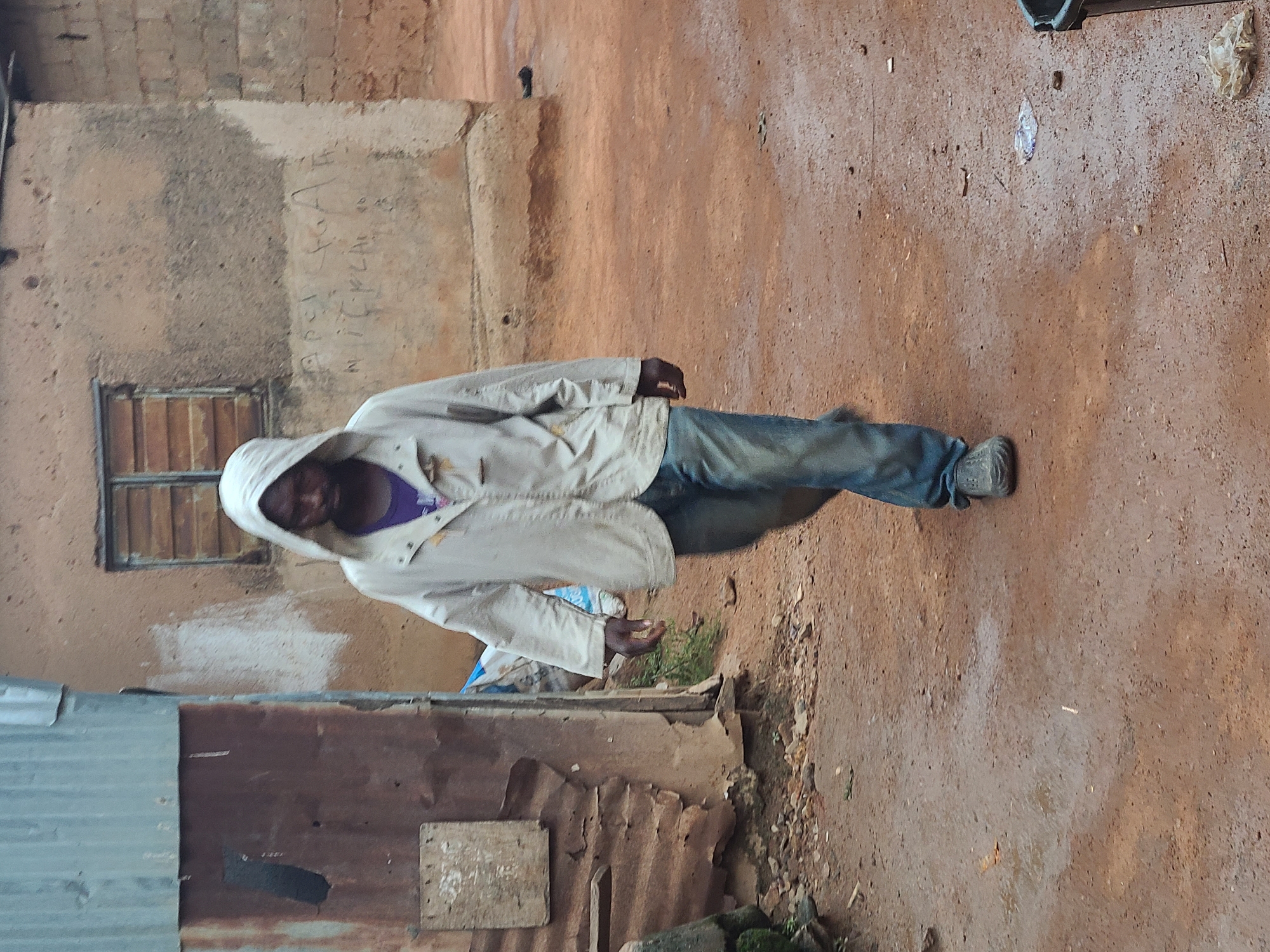 A man with dark skin and a purple shirt is wearing a light-colored hooded jacket and blue jeans. He is standing on a dirt ground next to a building with corrugated metal and a window. The background shows a dilapidated wall with faint writing. The location is likely in Jos, Nigeria.