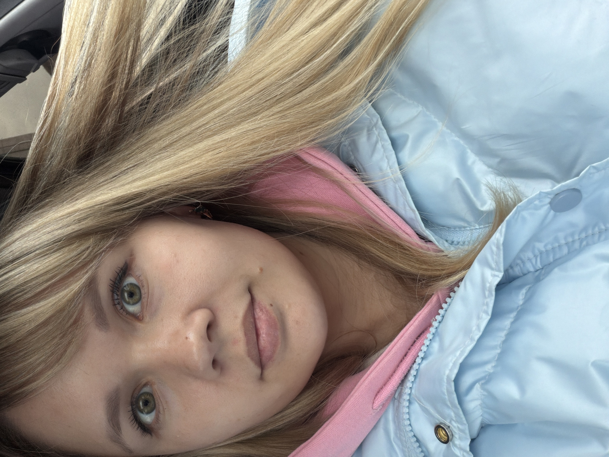 A close-up, portrait-style image features a young person, likely female, with long, straight, light blonde hair that cascades across the frame. The subject's head is tilted significantly to the left, with the image itself also rotated. She has striking green eyes, which are looking directly at the viewer. Her complexion appears clear, with a few subtle marks or freckles on her face. Her lips are slightly parted, revealing a neutral expression, possibly a relaxed or contemplative look.
She is dressed in a light blue puffer jacket, identifiable by its shiny, quilted fabric and visible zipper and snap buttons. Underneath the blue jacket, a vibrant pink hoodie is visible, peeking out around her neck and chest area. The hair dominates a large portion of the upper and left side of the frame, creating a soft, flowing texture. The background is mostly obscured by her hair and clothing, but dark, nondescript shapes in the upper left hint at an indoor setting, possibly inside a vehicle due to the texture and color suggesting a car's interior. The lighting is bright and even, suggesting daytime conditions. There is no visible text in the image.