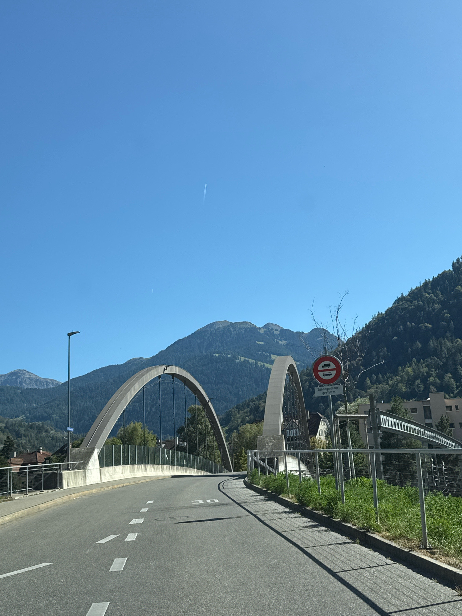 A clear, bright day is depicted as the image captures a curved road leading onto a modern bridge. The bridge has a distinctive arch design, with one arch appearing larger and more prominent than the other. The road is marked with white lines, and a speed limit sign indicating "30" is visible on the pavement. A metal railing lines the side of the road and bridge, separating it from a grassy area and a slope. In the background, lush green mountains rise sharply against a brilliant blue sky, with a few faint contrails from an aircraft visible. A streetlamp stands tall on the left side of the road. A traffic sign with a red circle and a horizontal line, depicting a truck with a red circle and line through it, and text beneath it indicating "Zubringerdienst gestattet," suggests restrictions on certain types of traffic. The scene appears to be in a scenic, mountainous region.