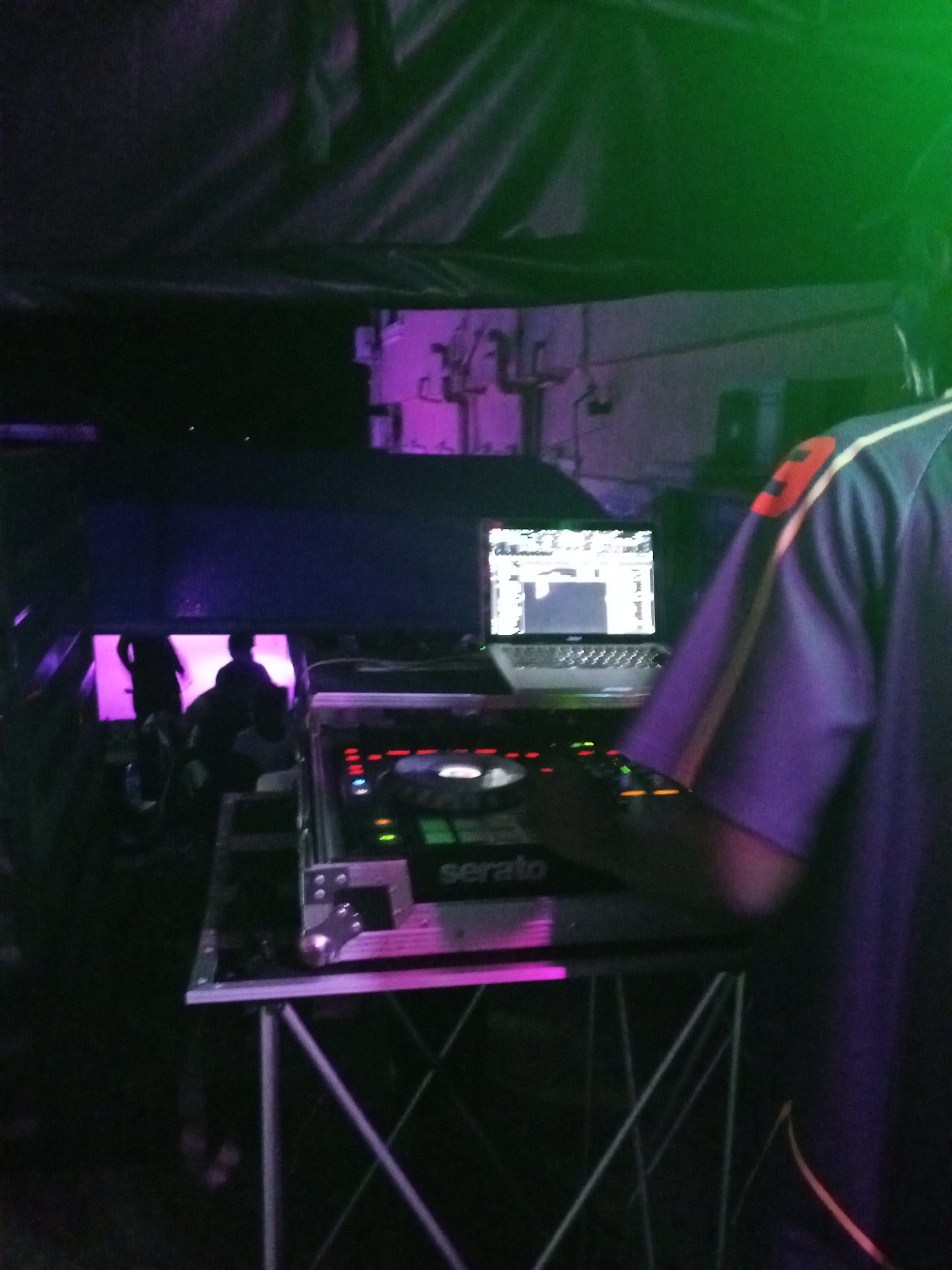 A person in a blue and white shirt is standing behind a DJ setup with a laptop and a Serato mixer, likely in a performance setting in Lokoja, Nigeria. Other people can be seen in the background bathed in a purple glow. The scene is dark.