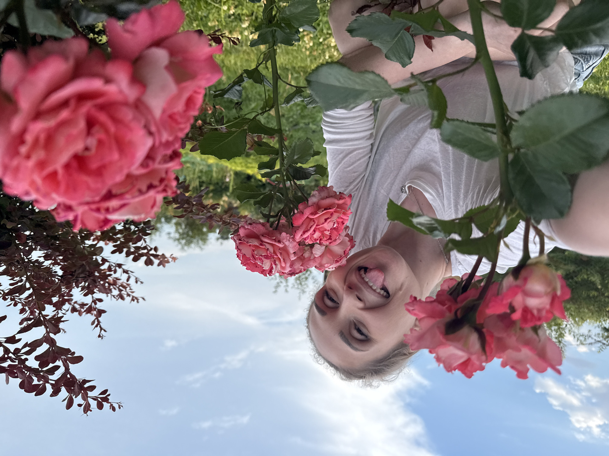 A young woman with blonde hair is pictured outdoors, with the sky and greenery in the background. She is making a playful face, sticking her tongue out and smiling broadly, with her eyes looking towards the camera. She is wearing a white t-shirt. Several pink roses are visible in the foreground, some in focus and some blurred, as well as branches with small, reddish-brown leaves. The image is oriented upside down. The lighting suggests it is daytime, possibly late afternoon or early evening, with a bright sky and some clouds. The scene appears to be a garden or natural outdoor setting, possibly in Chaikovsky, Russia, given the location context.