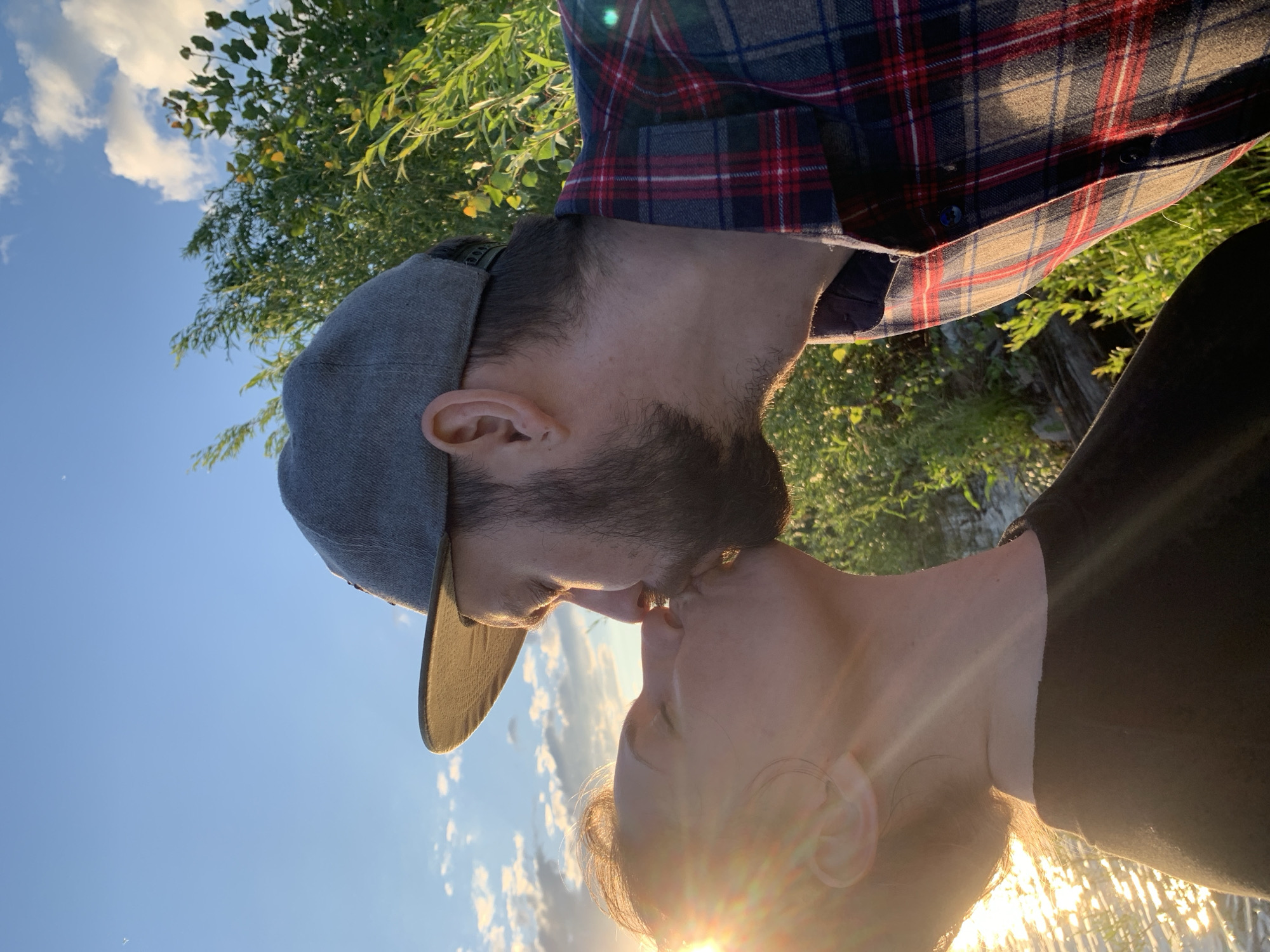 A couple is kissing in the golden hour sun, near the Kama River in Chaikovsky, Russia. The man is wearing a grey baseball cap and a red and blue plaid shirt. The woman is wearing a black shirt. The background is filled with lush green foliage and a bright blue sky with scattered clouds. The sun is setting, casting a warm glow on the scene and creating lens flare. The couple appears to be on a slight incline, with the river visible in the lower left corner of the image.