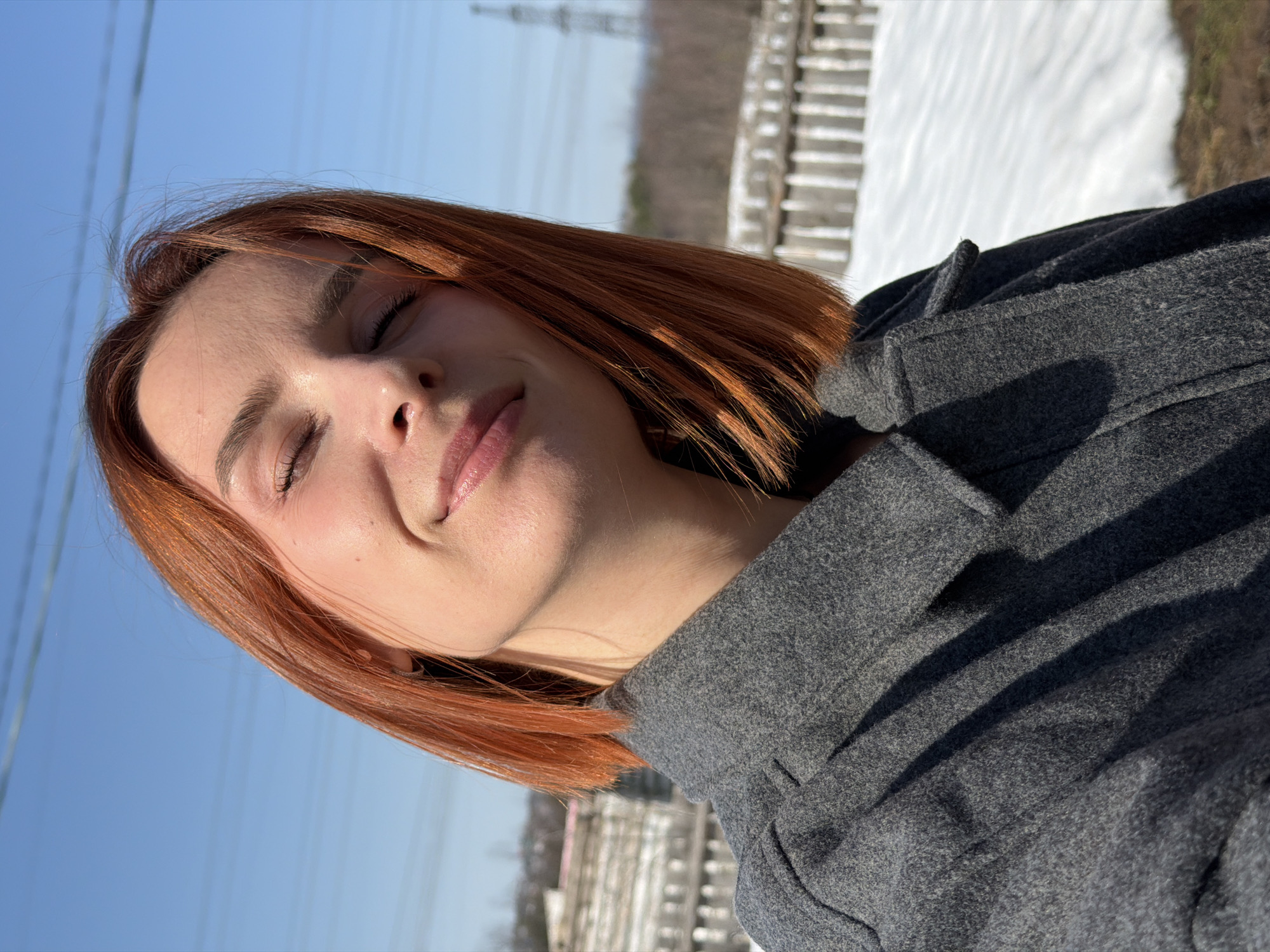 A portrait of a woman with red hair and a grey coat, eyes closed and smiling gently, in an outdoor environment. The background shows a bright blue sky with power lines visible and a patch of snow. The location is identified as Chaikovsky, Russia.