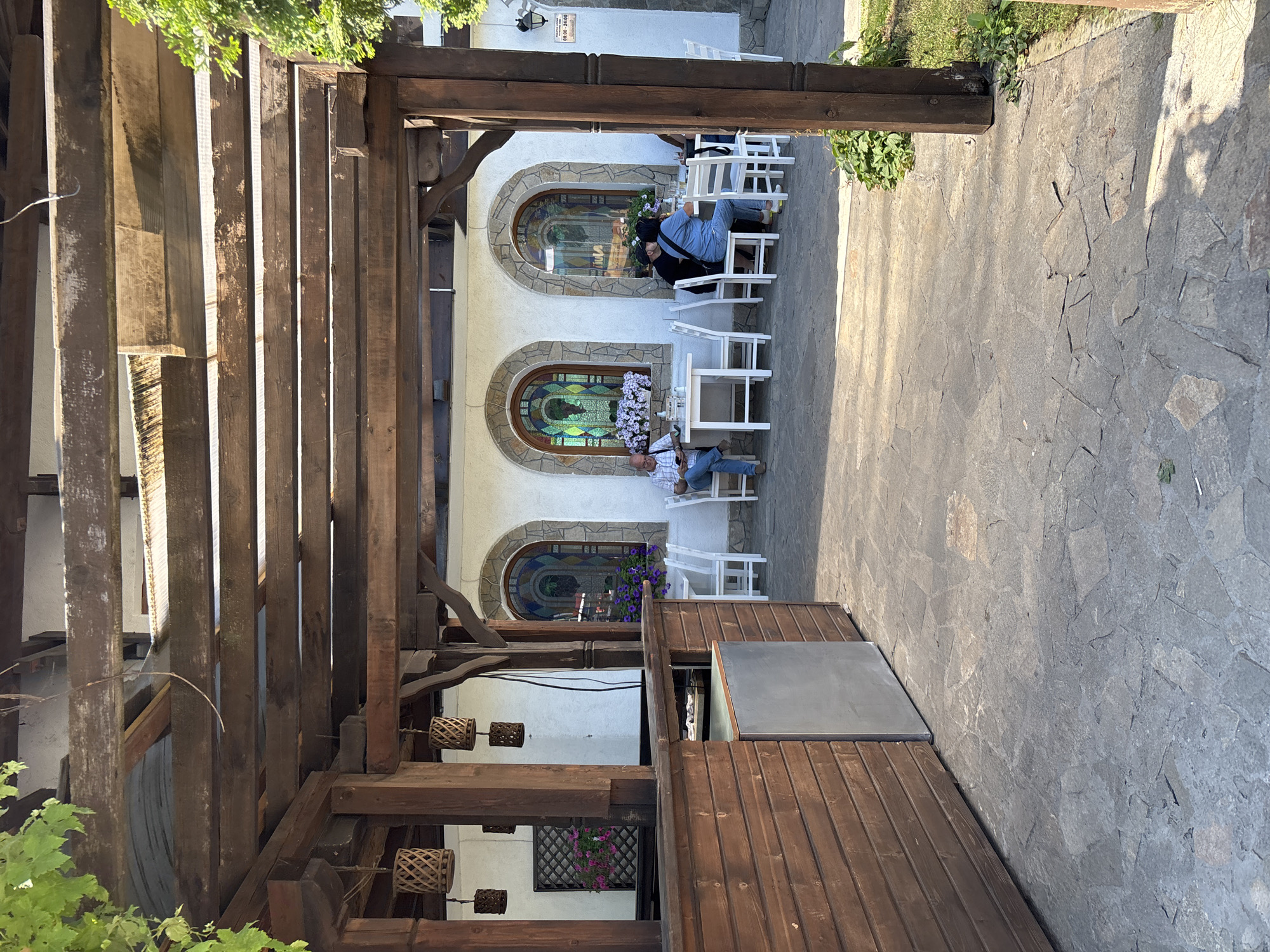 The image depicts an outdoor seating area with a wooden pergola structure in the foreground. Several people are seated at tables, appearing to be patrons of a restaurant or cafe. The building in the background features arched windows with stained glass and is painted white. Planters with flowers are visible near the windows. The ground is paved with irregular stone slabs. A sign is attached to the building's exterior, partially visible. The setting suggests a relaxed, possibly daytime, social gathering in a town or city. The location is identified as Nesebar, Bulgaria.