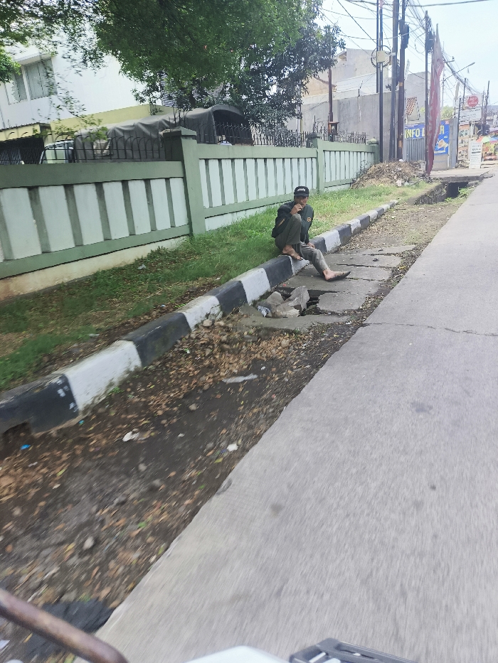 The image depicts a man sitting on a curb in a street located in Ngamprah, Indonesia (latitude: -6.865, longitude: 107.513). The man is wearing a dark green jacket and black pants. He is looking towards the left. He is sitting with his legs crossed. The curb has a black and white striped border. It is on the side of a paved road. The road is empty and it is covered in shadows. There is a green metal fence to the left of the curb, behind it there is a building with a tiled roof. The ground is covered in dry, brown leaves. There is a small, rectangular pit in the sidewalk near the man. The scene appears to be taken from a vehicle moving on the road.