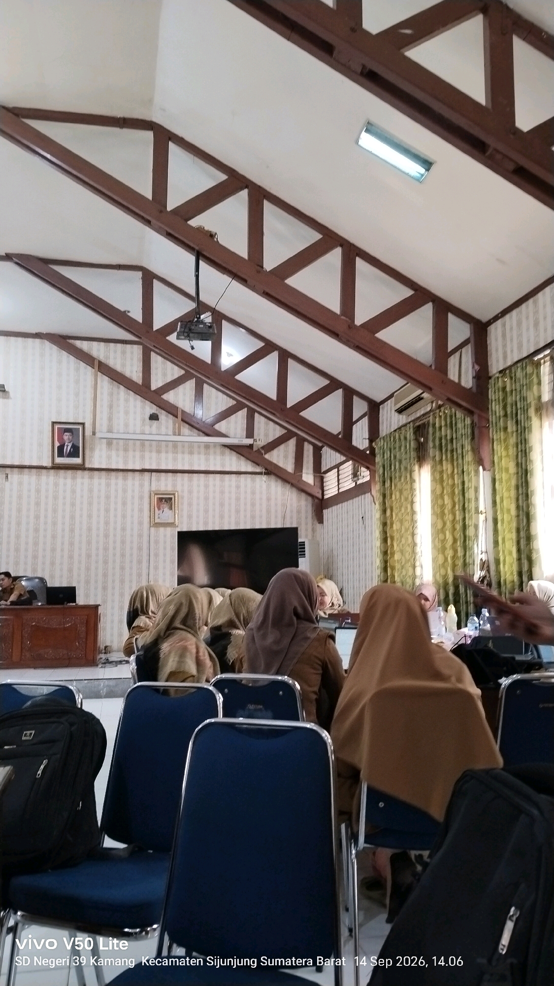 The image depicts an indoor scene, likely a conference room or classroom, within the building of SD Negeri 39 Kamang, located in the Sijunjung Regency of West Sumatra, Indonesia. The photograph was taken on September 14, 2026, at 2:06 PM.
The room features a distinctive beamed ceiling with exposed wooden rafters. A projector is mounted on the ceiling, positioned above a large flat-screen television screen. The walls are adorned with patterned wallpaper. Two framed portraits are visible on the wall, one of which appears to be a formal portrait of a man in uniform.
In the foreground, several dark blue chairs with metal frames are arranged in rows. One chair on the left has a black backpack placed on it. A wooden podium or desk is visible to the left of the frame.
Several individuals are seated, mostly facing away from the camera. They are dressed in modest attire, with many wearing headscarves. Their posture suggests they are attending a presentation or meeting. The lighting in the room appears to be a mix of natural light from the windows, which are covered by green curtains, and artificial lighting from a fluorescent fixture on the ceiling. The overall atmosphere is that of a formal gathering or educational event.