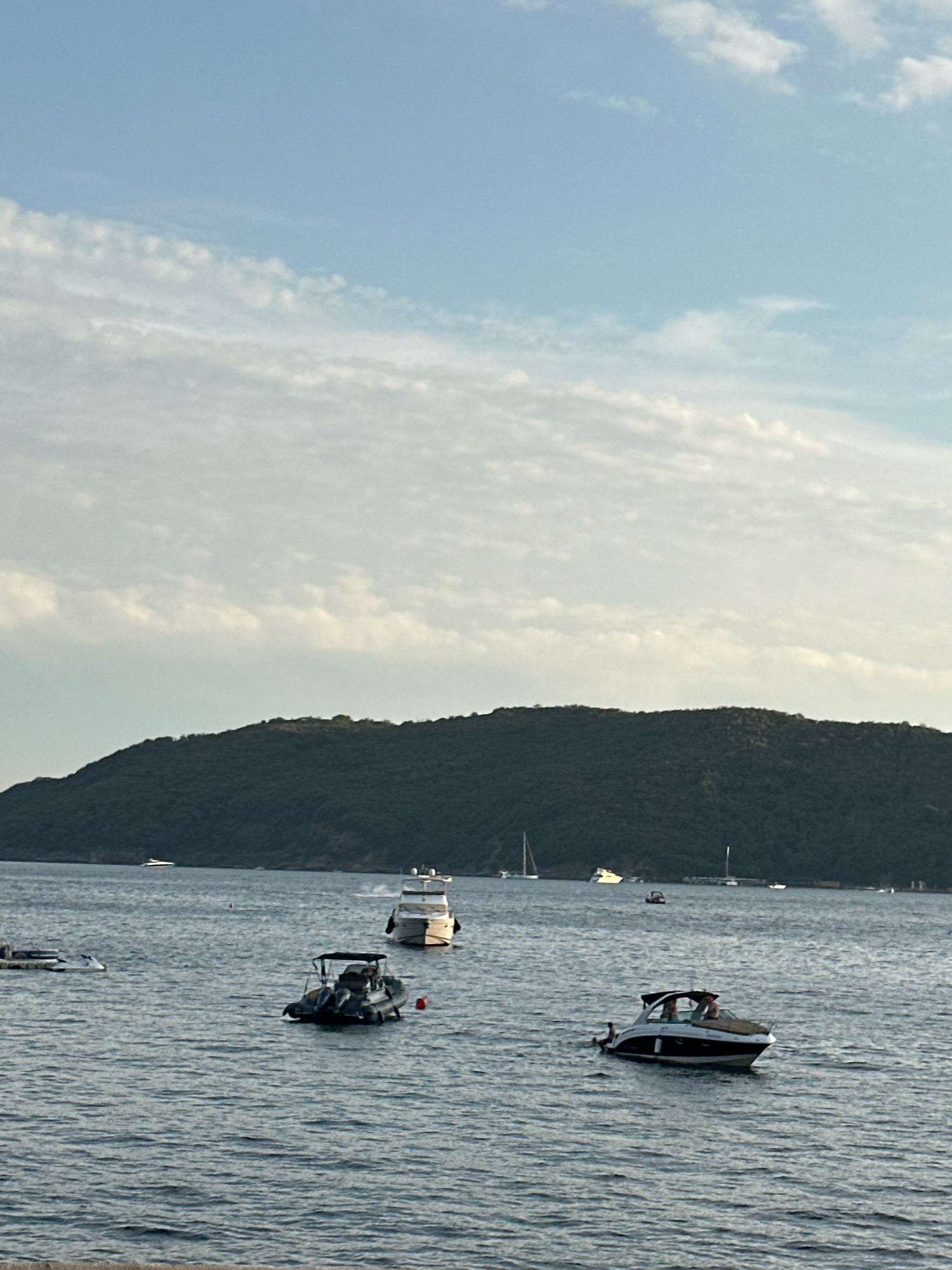 The image depicts a scenic view of the Adriatic Sea near Budva, Montenegro. In the foreground, the deep blue water is rippled, suggesting a gentle breeze. Several boats are visible on the water. To the right, a sleek, white and black motorboat with a canopy is cruising, carrying at least two people. To the left of this boat, a dark-colored inflatable boat with a canopy is stationary, with what appears to be a person sitting in the stern. Further in the distance, a larger white motor yacht is moving towards the left. On the far left, another smaller boat or watercraft is visible. In the background, a large, densely forested island or peninsula dominates the horizon, its dark green slopes meeting the light blue sky. The sky itself is adorned with scattered, wispy white clouds, particularly concentrated in the upper half of the image, with a clearer blue expanse towards the top. A few distant sailboats are also faintly visible on the water, adding to the nautical atmosphere. The overall impression is a tranquil summer day at sea.