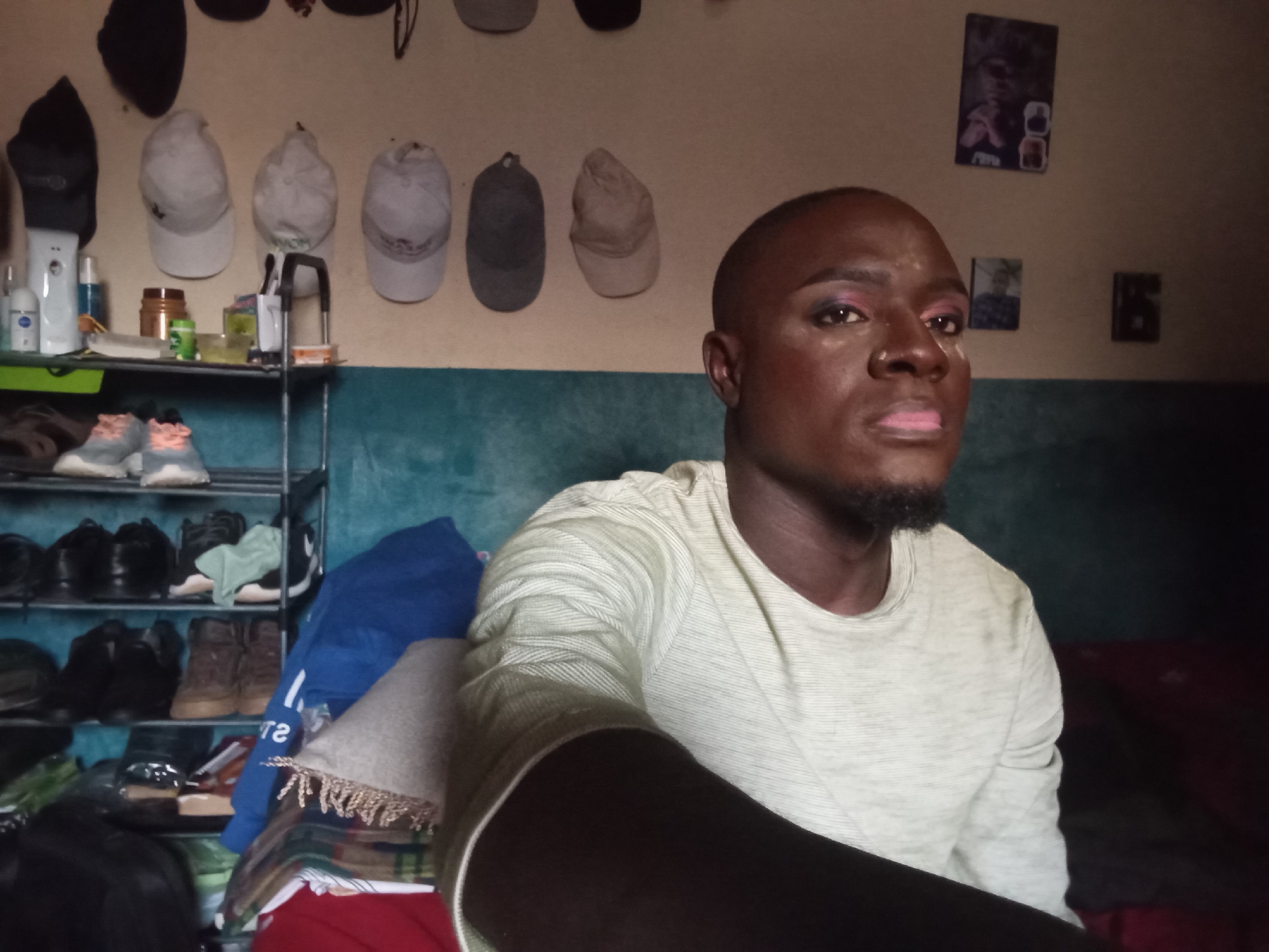 This is a head and shoulders selfie of a Black individual with makeup applied to their face, including eyeshadow and lipstick. They are wearing a light-colored, long-sleeved t-shirt. In the background, a shelf unit holds various items, including shoes, hats, and bottles. The wall behind the shelf unit is decorated with framed pictures. The overall environment appears to be an indoor residential setting in Rukuba Road, Nigeria.