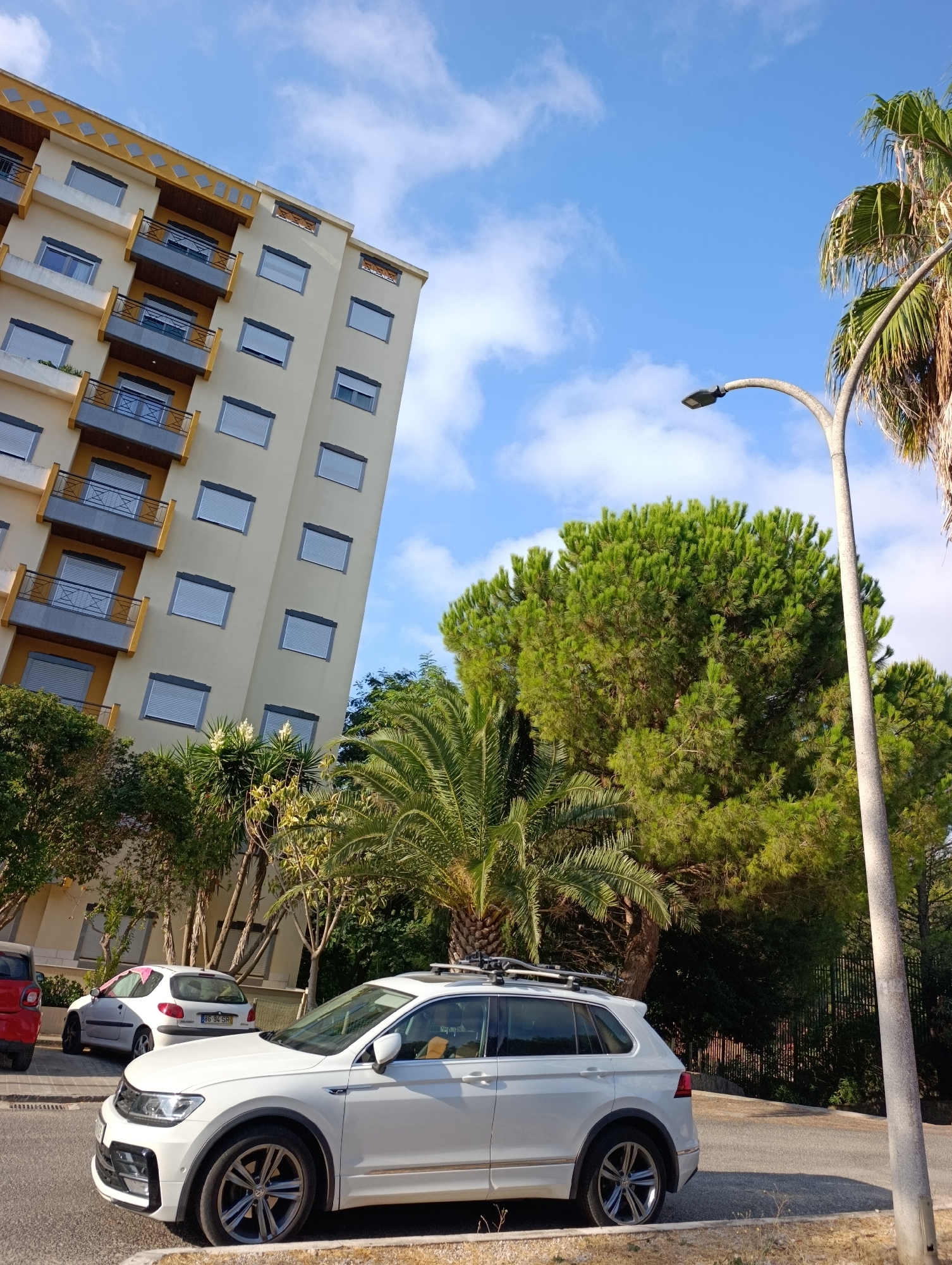 In the city of Setúbal, Portugal, on a clear day with scattered clouds, a white Volkswagen Tiguan SUV is parked on the right side of a paved road. The car features a roof rack and is positioned in front of a building and a dense group of trees. To the left of the Tiguan, a white Peugeot 206 hatchback is also parked, with a pink fabric draped over its rear windshield. Further left, the red rear of a Smart car is visible. Behind the parked cars, a multi-story building with cream-colored walls and yellow accents on its balconies is visible. The building has numerous windows, many with closed grey blinds. A tall lamppost with a modern design curves over the road towards the right side of the frame. The scene is framed by various types of trees, including palm trees and a large, lush green pine tree. The overall setting suggests a residential or commercial area with abundant vegetation, typical of a Mediterranean climate.