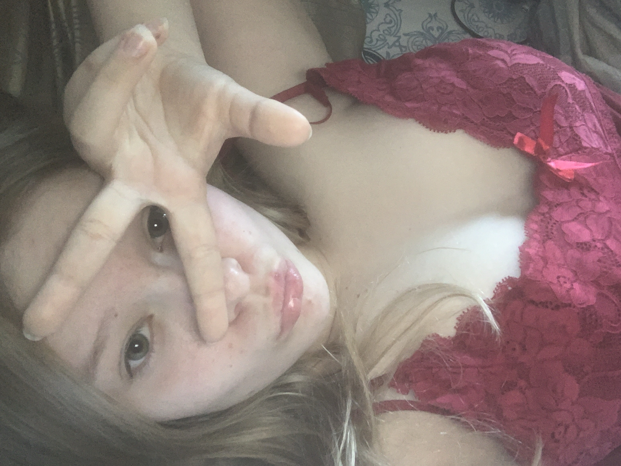 A young woman is lying down, looking directly at the camera. She is holding her hand up in a "peace" sign, partially obscuring her face. She is wearing a red, lacy top with a bow. The background is blurry and indistinct. There is no evidence to suggest the location is Saint Petersburg, Russia.