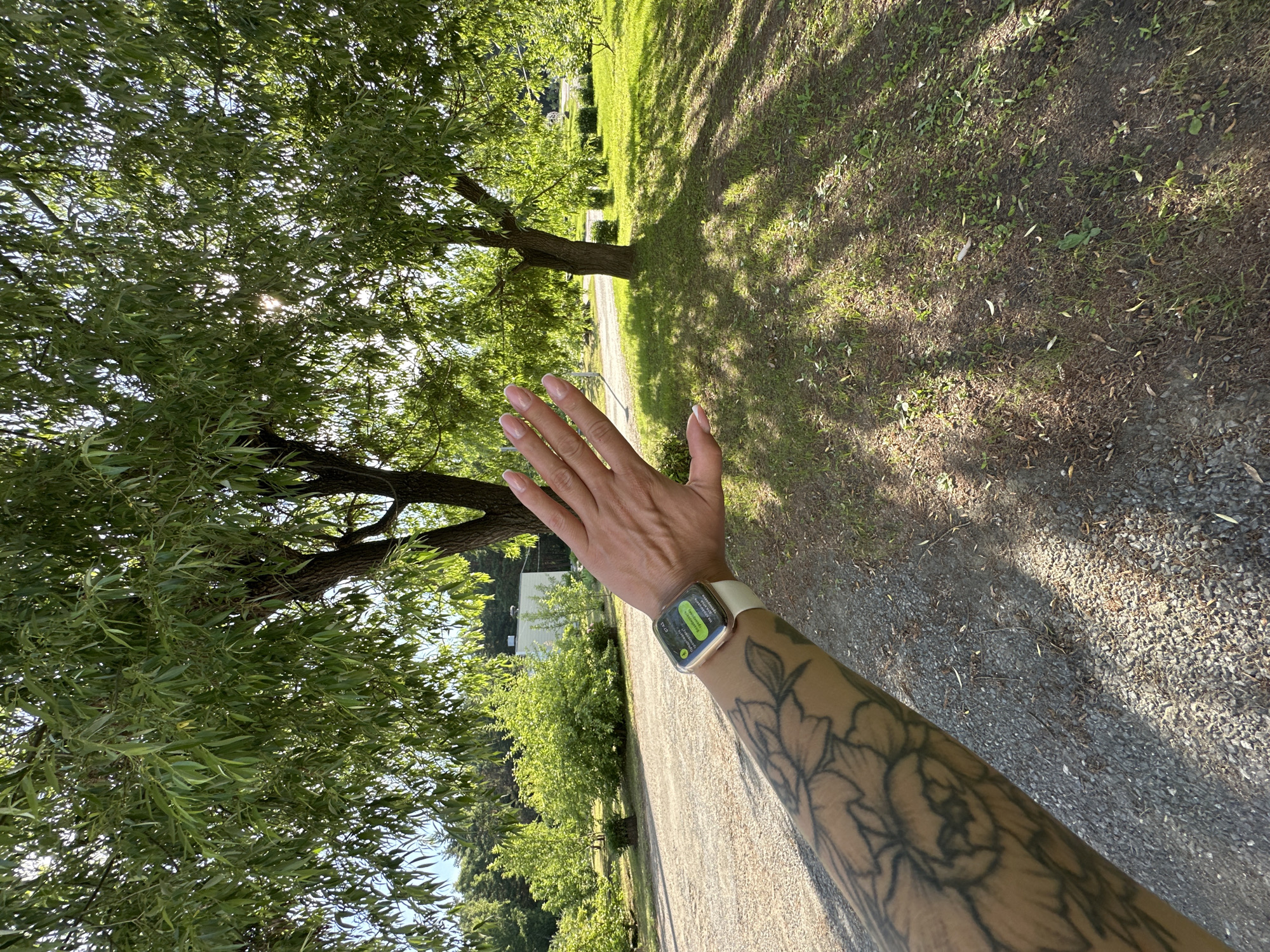 A person's arm, adorned with a floral tattoo, extends towards the viewer, partially obscuring a smartwatch on their wrist. The smartwatch displays text in Russian, including the time "07:43." The arm is positioned in front of a backdrop of lush green trees, casting dappled shadows on a gravel pathway and a grassy area. In the distance, a building with a white facade is visible amidst more greenery. The overall scene suggests an outdoor setting, possibly a park or rural area, with sunlight filtering through the leaves of the trees. The location is identified as Duvankul', Russia.