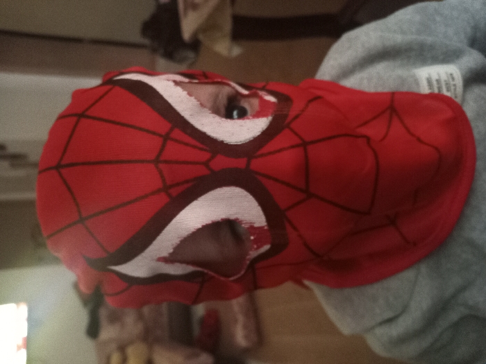 An individual, likely a child, is depicted in a medium close-up, facing directly towards the camera. The person is wearing a red fabric mask designed to resemble Spider-Man, featuring black web patterns and white eye sections outlined in dark red. The mask covers the entirety of the head and neck, with only the subject's eyes visible through irregular openings. Below the mask, a grey hooded sweatshirt is partially visible, with a white textile label displaying black text on the right chest area. The background is an out-of-focus indoor environment, presenting a light-colored sofa or armchair to the left and indistinct furniture or objects to the right. A soft, diffused light source is present in the upper left background. The scene is located in a residential or interior space within Dar Bouazza, Morocco.