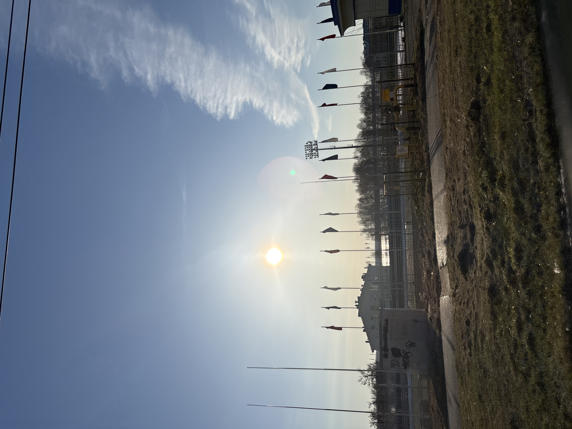 This is an outdoor, horizontally oriented image captured on a bright, sunny day, likely in Votkinsk, Russia. The sky dominates the upper portion of the frame, displaying a clear, light blue hue with a prominent band of wispy, white clouds (likely cirrus) extending from the upper left to the upper right.
The sun is a brilliant orb in the middle-left of the image, casting a strong glare and lens flare. Below the sun and clouds, a line of tall flagpoles extends diagonally from the middle ground towards the background, each flying a small, rectangular flag. The flags vary in color, with some appearing red, others white or light-colored, and some dark.
Along the lower half of the image, a dirt or gravel path runs parallel to a sturdy metal fence. Patches of dry, light green grass are visible on either side of the path. Behind the fence, several structures and bare trees form the background. Notably, on a building or structure situated to the right of the sun and below some flags, the Cyrillic text "СТАДИОН" (STADIUM) is clearly visible, identifying the location as a stadium. Further along the fence line, a large stadium light array or scoreboard can be seen, its display unreadable from this distance.
In the far background, additional buildings and a line of bare trees are present, suggesting either late autumn or early spring. On a concrete wall in the bottom right corner, some graffiti is visible. Power lines run vertically in the far upper left. No people are present in the scene. The weather appears clear and calm.