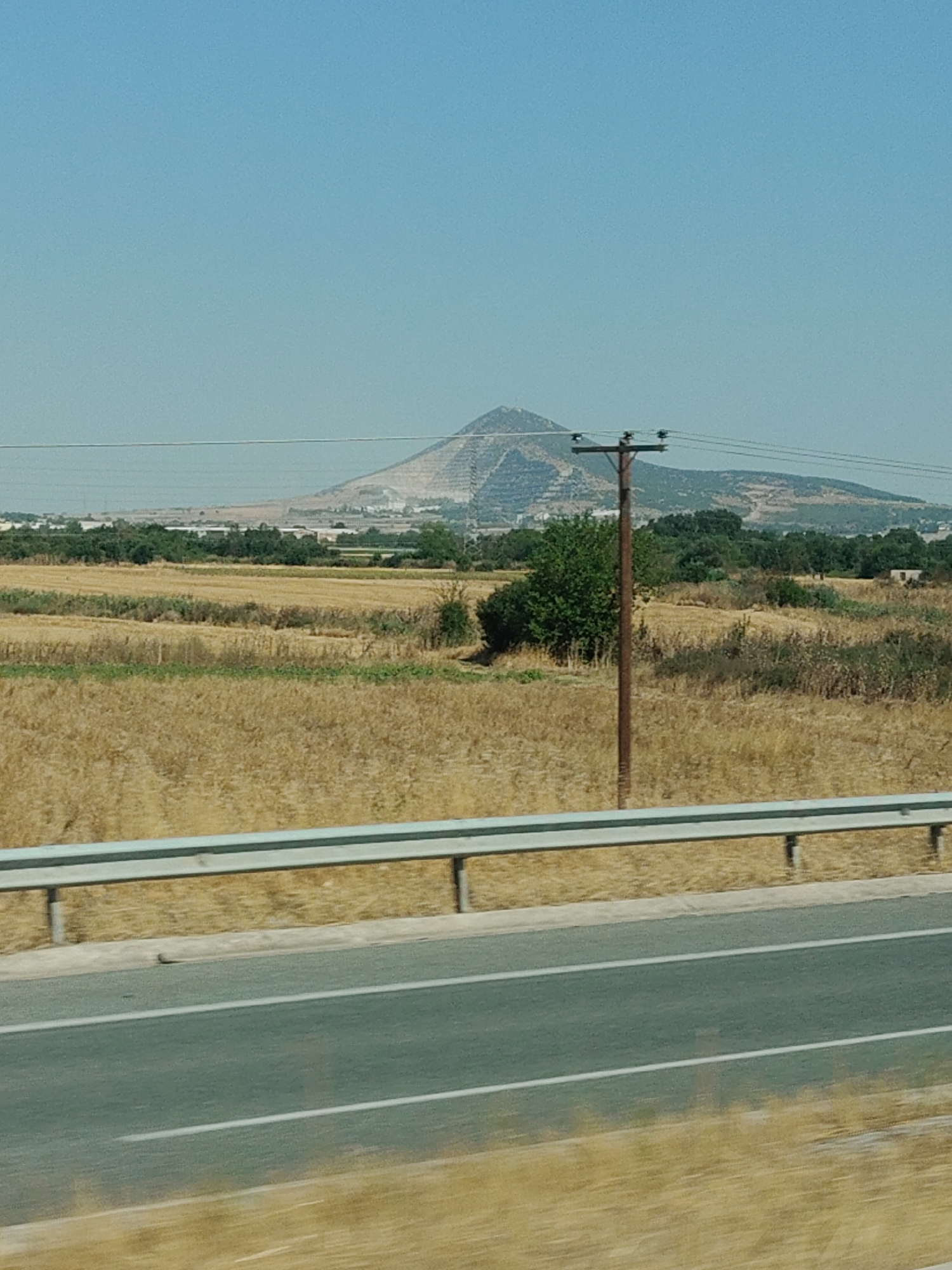 The image shows a wide shot of a rural landscape under a clear blue sky. In the foreground, a highway is visible with a metallic guardrail separating it from a dry, yellowish field. Beyond the field, there are scattered trees and a single wooden utility pole with wires. In the background, a large, tiered mound dominates the horizon. This mound appears to be man-made and shows signs of excavation or construction, with lighter-colored material visible on its slopes. The surrounding area also appears to have some scattered buildings and vegetation. The location is identified as Mygdonia Municipal Unit, Greece.