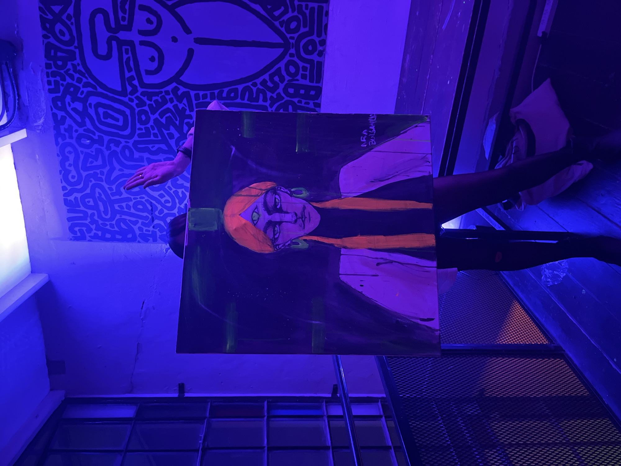 The image, heavily bathed in blue light, depicts a person holding a painting in what appears to be an indoor space, potentially an art gallery or studio in Buenos Aires, Argentina. The artwork on the painting shows a stylized figure with orange hair, a headdress, and green eyes, painted against a dark background. The signature "ARA BOBALANO" is visible. To the left, a wall mural features black and white abstract patterns. The person holding the painting is partially obscured, but their hands are visible. The floor is made of wood. A metal grate and window are situated below and to the right. The bright blue light affects color perception, making it difficult to determine the exact hues of the artwork and surrounding objects. The environment feels creative and unconventional.