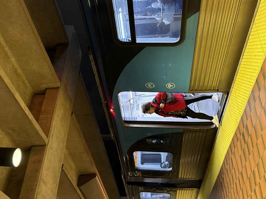 A train car, oriented vertically, shows a woman in a red jacket entering or exiting. Inside, people are reflected in the windows. Outside, a textured yellow platform edge is visible. Adjacent to the train is a light-colored concrete structure with visible beams and a light fixture. The image is taken at an angle.