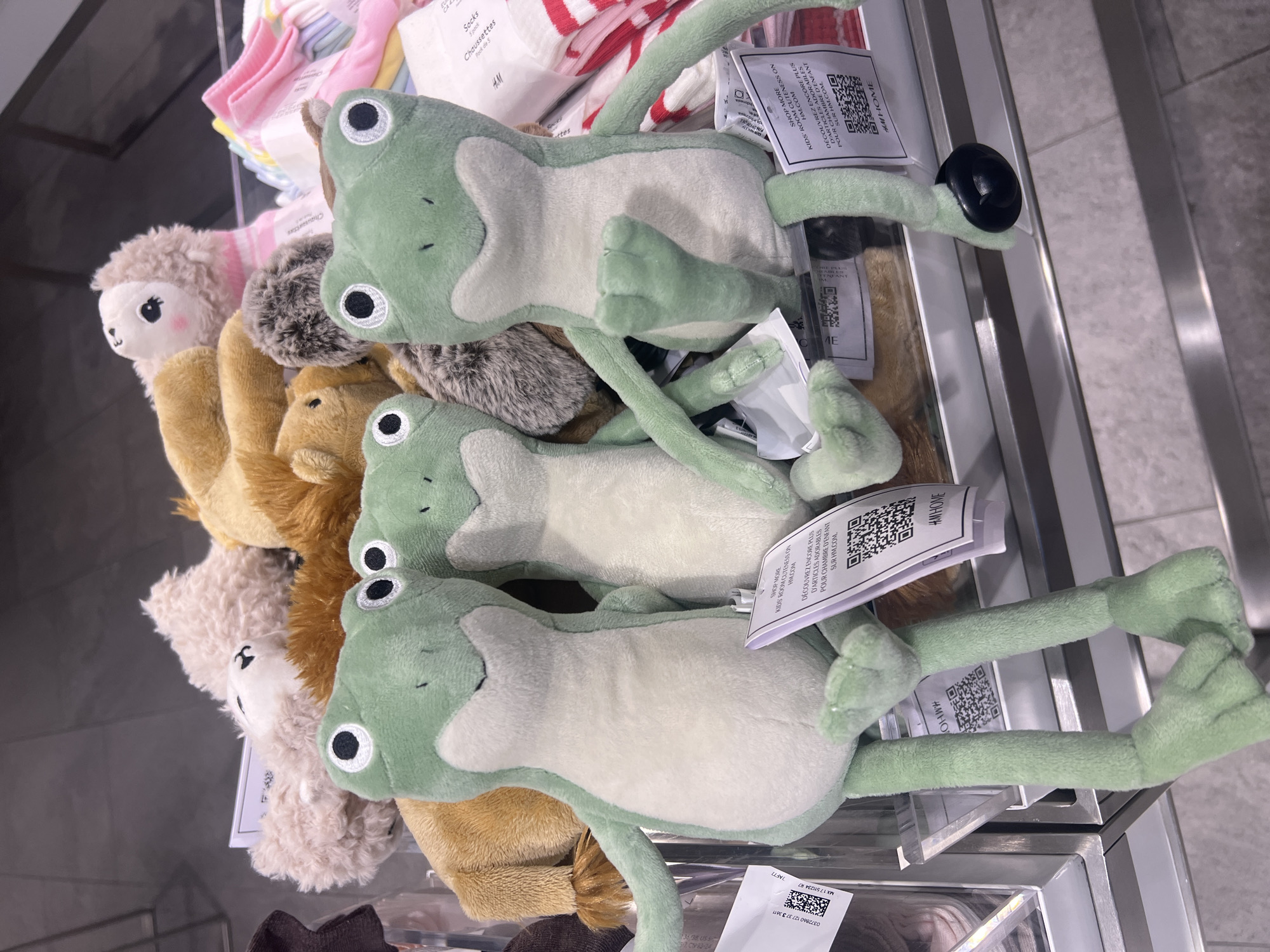 A shelf displays a variety of stuffed animals and infant apparel. Three green frog plush toys dominate the upper right, each bearing a white tag with black print and a QR code. To the left of the frogs are several smaller, indistinct stuffed animals in beige, brown and cream colors. At the top of the frame are stacked pink and white infant socks. The scene appears to be inside of a retail store. There is no evidence to support a connection to the city of San Pedro Cholula, Mexico.