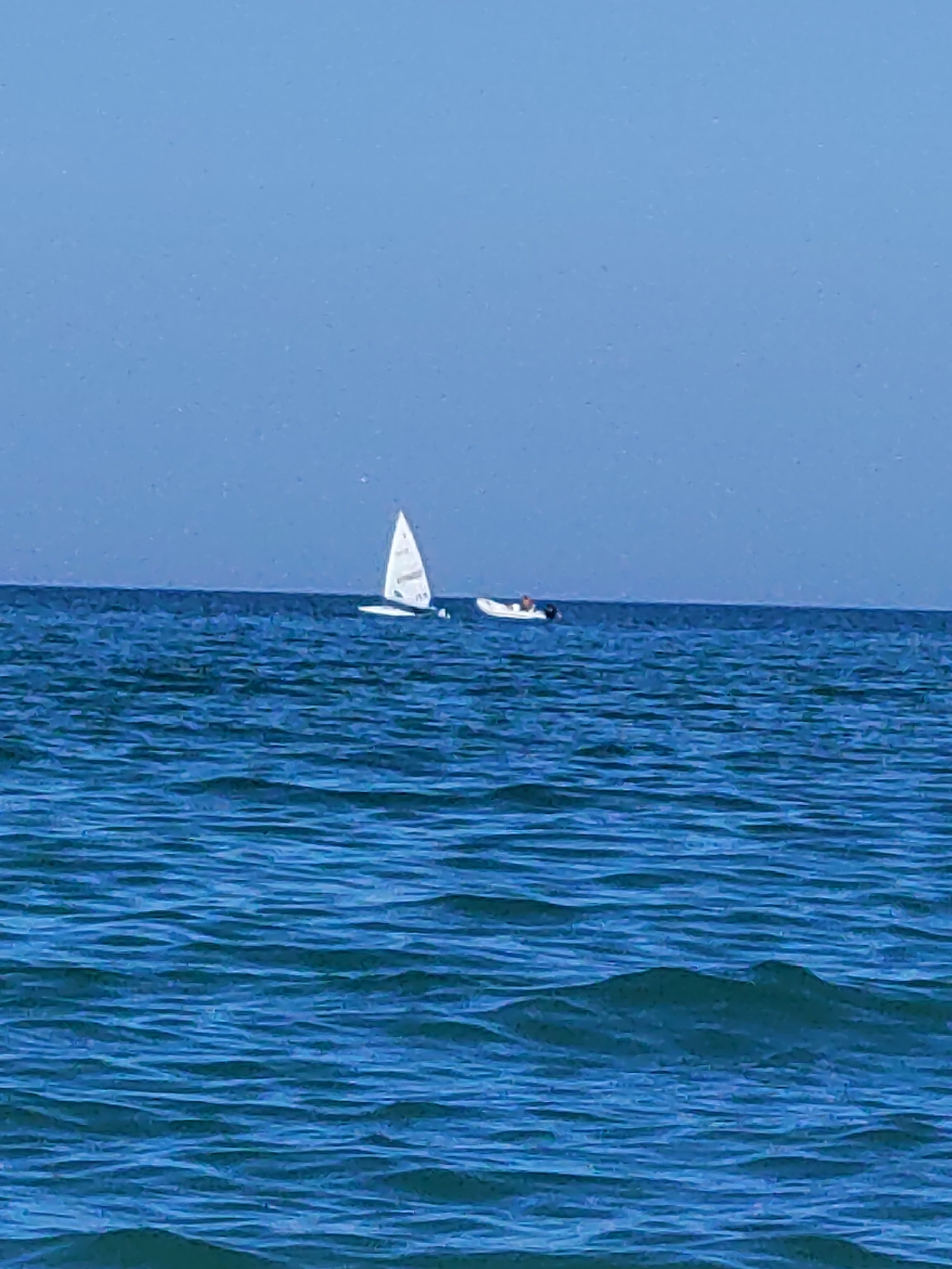In the open sea off the coast of Porto Recanati, Italy, a white sailboat with a single sail is visible. To the right of the sailboat, a small white inflatable boat with an outboard motor is also present. The sea is calm, with gentle waves, and the sky is a clear, light blue. The horizon line is distinct, separating the water from the sky.