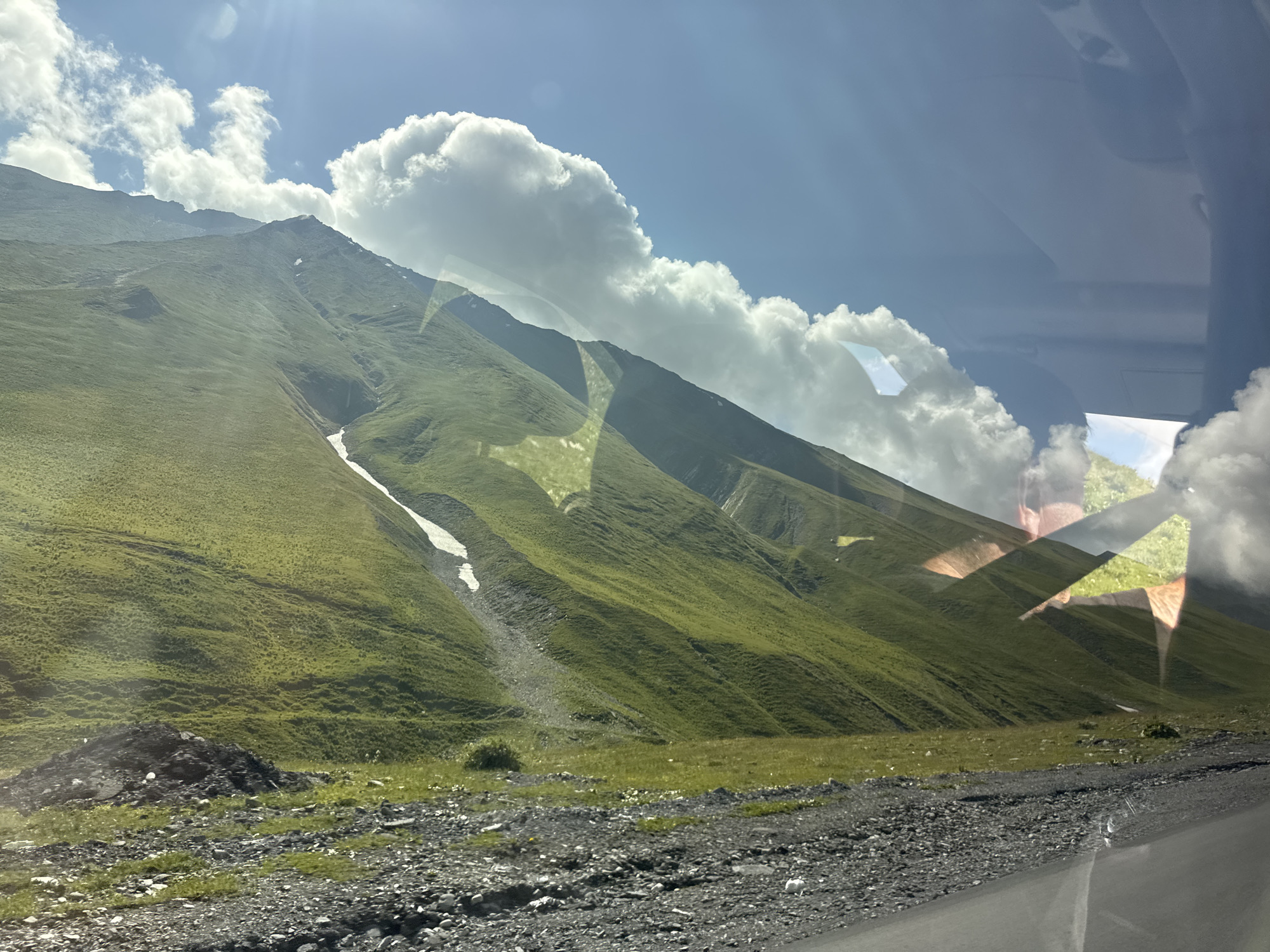 A wide, green, mountainous landscape is seen under a bright, partly cloudy sky. The mountainside is covered in lush green grass with patches of snow, particularly visible on a steep slope on the left side of the frame. A gravelly road or path runs along the bottom of the image, leading into the foreground. The scene appears to be viewed from within a vehicle, as evidenced by reflections on the right side of the image, showing parts of what appears to be a car interior, as well as reflections of trees and sky on the glass. The reflections are somewhat distorted and abstract. This is a natural landscape, and it is not possible to identify a specific city location within Almasiani, Georgia from the provided image.