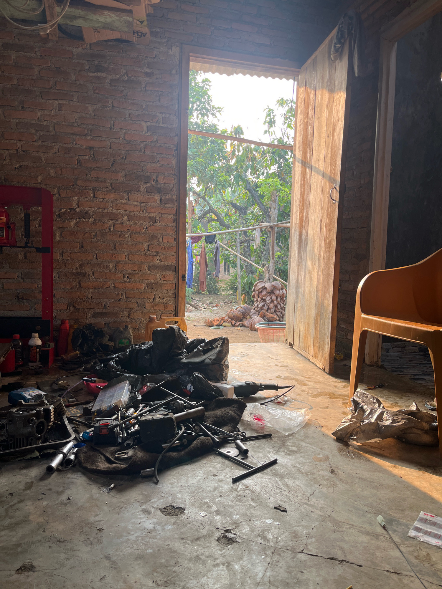 The image depicts the interior of a cluttered workshop or storage area, possibly located in Kebumen, Indonesia, with an open doorway leading to an outdoor scene. The workshop floor is concrete and dusty, littered with various tools, engine parts, black plastic bags, and containers of liquids. A red mechanical press stands against the brick wall on the left. In the foreground, a collection of tools and engine components are spread out on a dark cloth.
The doorway reveals a bright outdoor environment with lush green trees and foliage. Clothes are hanging on a line in the background, and a stack of coconuts is visible. A simple wooden door is open, allowing sunlight to stream into the workshop, casting long shadows. To the right, a portion of an orange plastic chair is visible. The overall impression is one of a functional, yet unorganized, workspace.