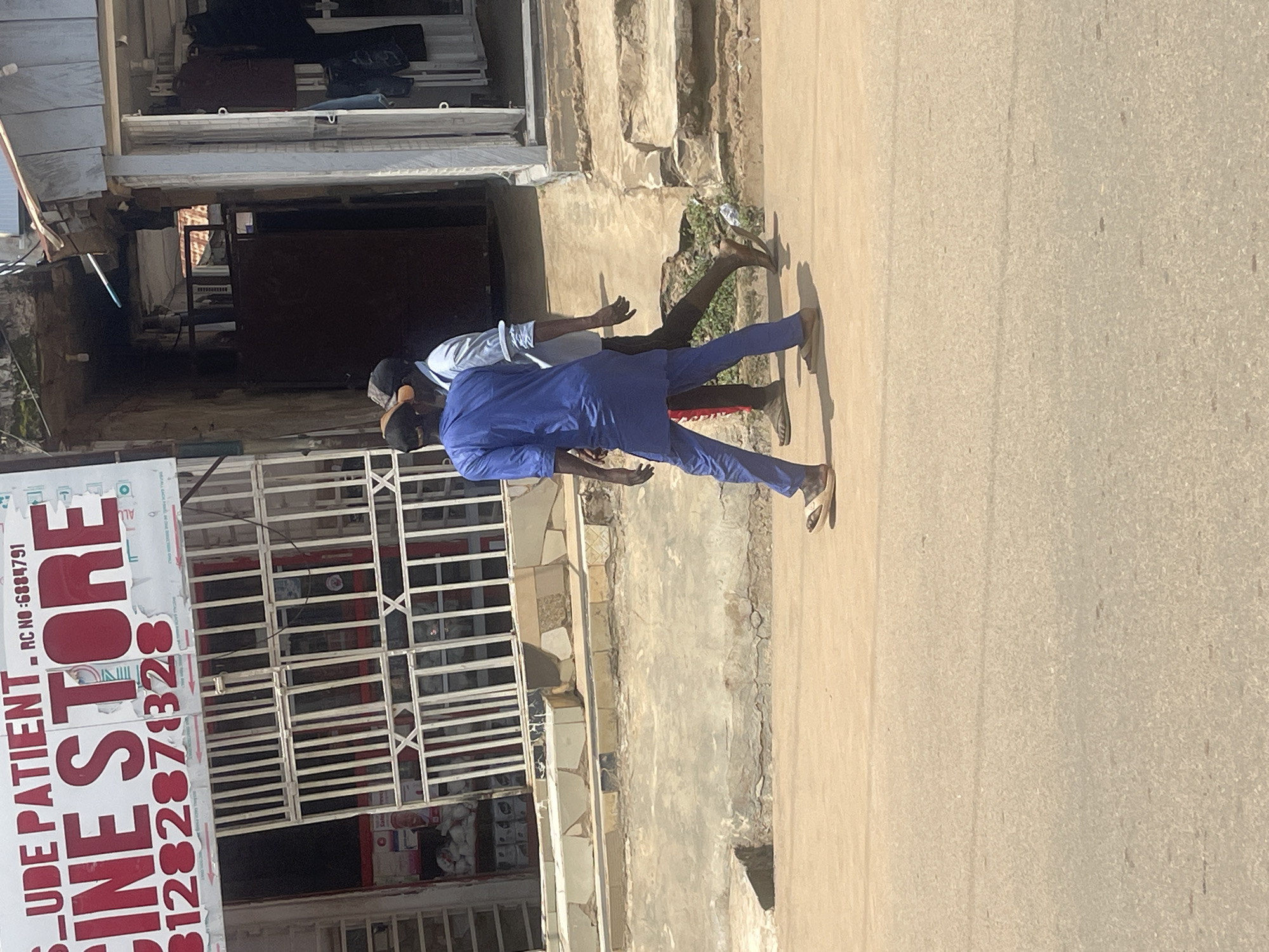The image shows a street scene in Abuja, Nigeria, focusing on the front of a business establishment. A prominent sign above the entrance reads "CINE STORE" in large red letters, with smaller text above it including "UDE PATIENT" and "RC NO: 6884791". Below the "CINE STORE" text is a phone number, "1282878328". The entrance itself is a metal gate with a white grill structure.
Two men are walking away from the camera on the street in front of the store. The man in the foreground is wearing a blue traditional outfit and sandals, and the man behind him is wearing a light blue shirt and dark pants, also in sandals. They are walking on a dusty, paved road. The surrounding environment includes a raised concrete sidewalk with textured tiles, and parts of buildings with various architectural styles. One building on the right appears to be a shop with clothes displayed. The overall lighting suggests it is daytime with sunlight casting shadows.