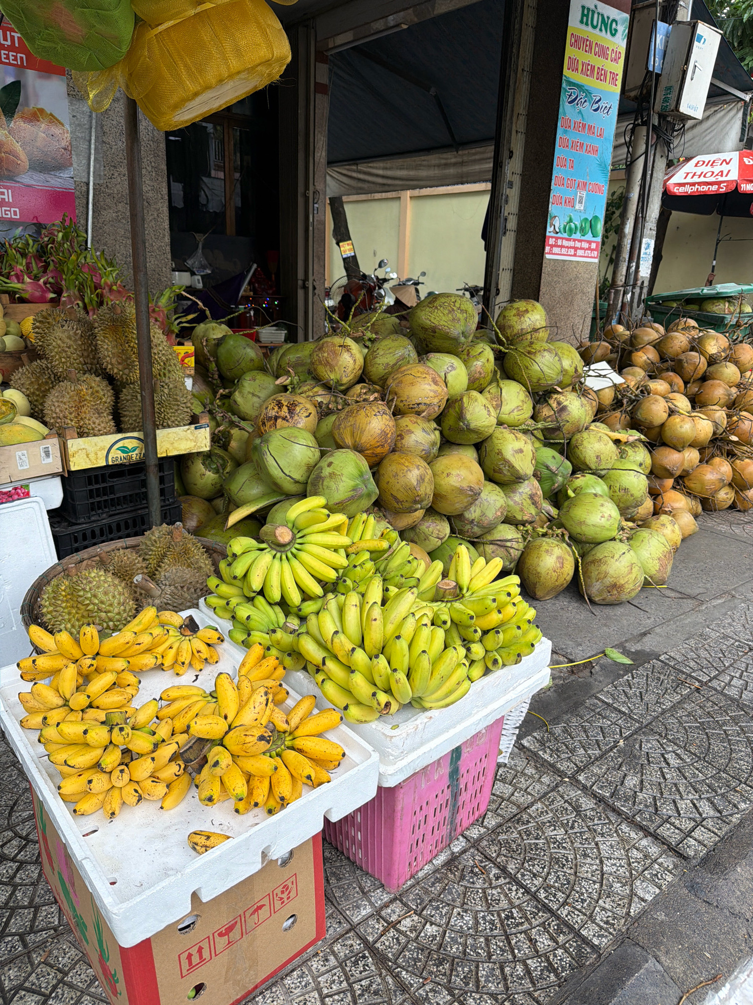 A fruit stand on a sidewalk in Da Nang, Vietnam, displays a variety of fresh produce. In the foreground, large piles of yellow bananas are arranged on white styrofoam boxes. Behind the bananas, a woven basket holds durian fruit, and to the left, more durian is stacked in wooden crates. Further back, a large mound of green coconuts is visible, with a pile of brown coconuts behind them. Overhead, two decorative woven hats, one yellow and one green, hang from a pole. A tall sign with Vietnamese text is mounted on a pole to the right of the fruit. The sidewalk is paved with patterned grey tiles, and a curb is visible. The background shows the entrance to a shop or building with a dark interior and some electrical wiring on the pole. A motorcycle is parked to the left of the shop entrance.