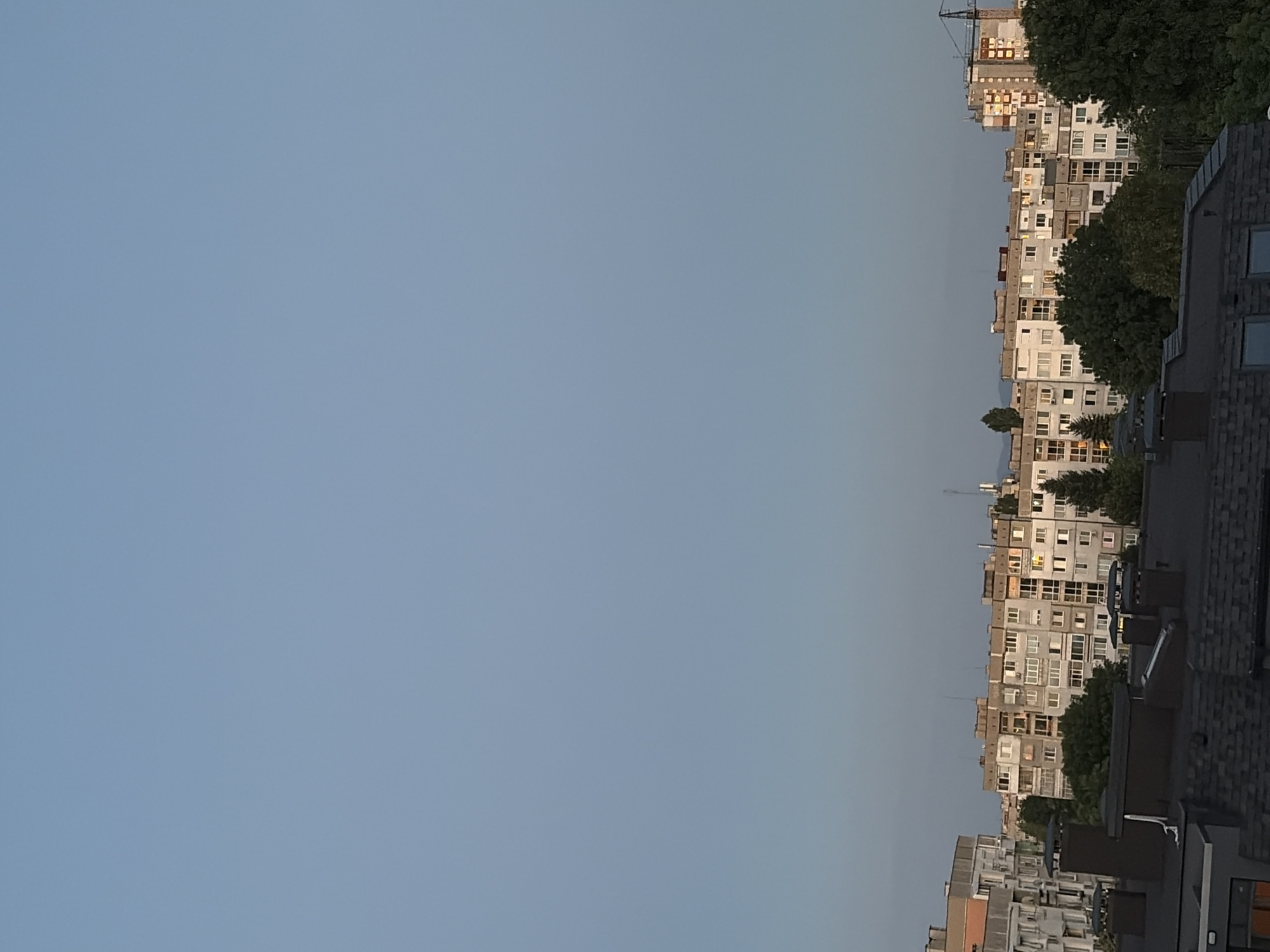 The image captures an urban skyline dominated by a large, light blue sky with no visible clouds. In the lower portion of the frame, a dense arrangement of multi-story apartment buildings is visible. These buildings are constructed in a uniform, utilitarian style, characterized by repetitive window patterns and flat roofs. Many of these structures appear to be quite old, with visible signs of wear and possibly dated architectural elements.
The apartment blocks are interspersed with several tall, mature trees, some of which are quite lush and green, while others show signs of seasonal change with sparser foliage. Several prominent chimneys, made of dark brick or stone, stand out on the rooftops in the foreground. One building further in the background has a construction crane situated next to it, suggesting ongoing development or renovation in the area. The light suggests it is either early morning or late evening, with a warm glow illuminating some of the building facades. No people or vehicles are discernible in the image. The overall impression is a residential area within a city, possibly in Eastern Europe given the architectural style of the buildings.