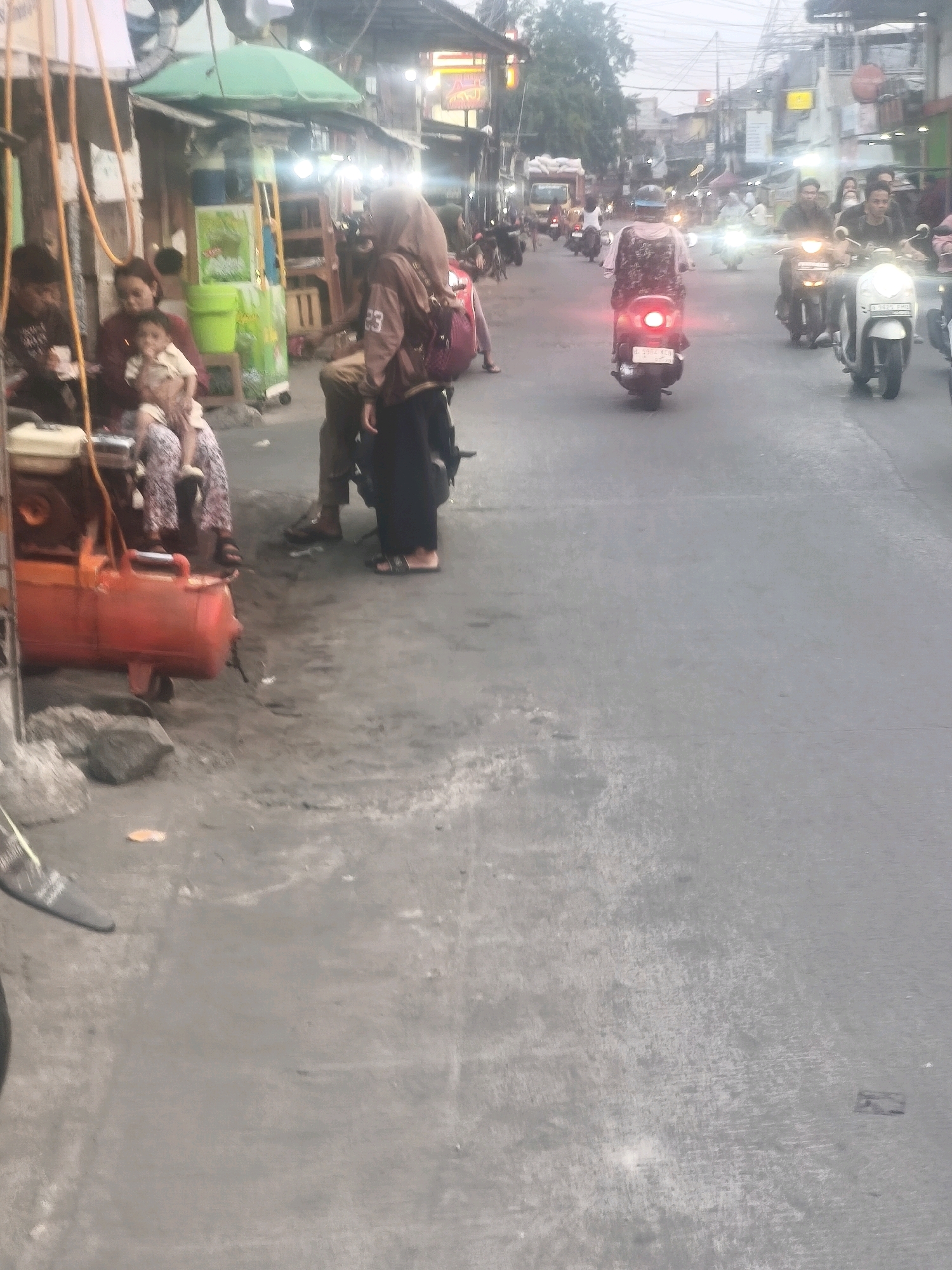 The image depicts a street scene in Bekasi, Indonesia, likely in the afternoon or early evening, with a mix of people and vehicles. In the foreground, to the left, is a bright orange air compressor with visible hoses. A group of people are seated and standing near a building with stalls and an umbrella. One woman, wearing a headscarf and a brown jacket with the number '23' on it, is standing and looking towards the street. She has a backpack on. Nearby, another woman is holding a child. To the right, the street is active with motorcycles. Several motorcycles are visible, some with riders. One white motorcycle is prominently featured in the middle ground, and further down the street, more motorcycles are moving. The road surface appears to be asphalt, and there are buildings lining the street, some with illuminated signs. The overall atmosphere suggests a busy urban environment.