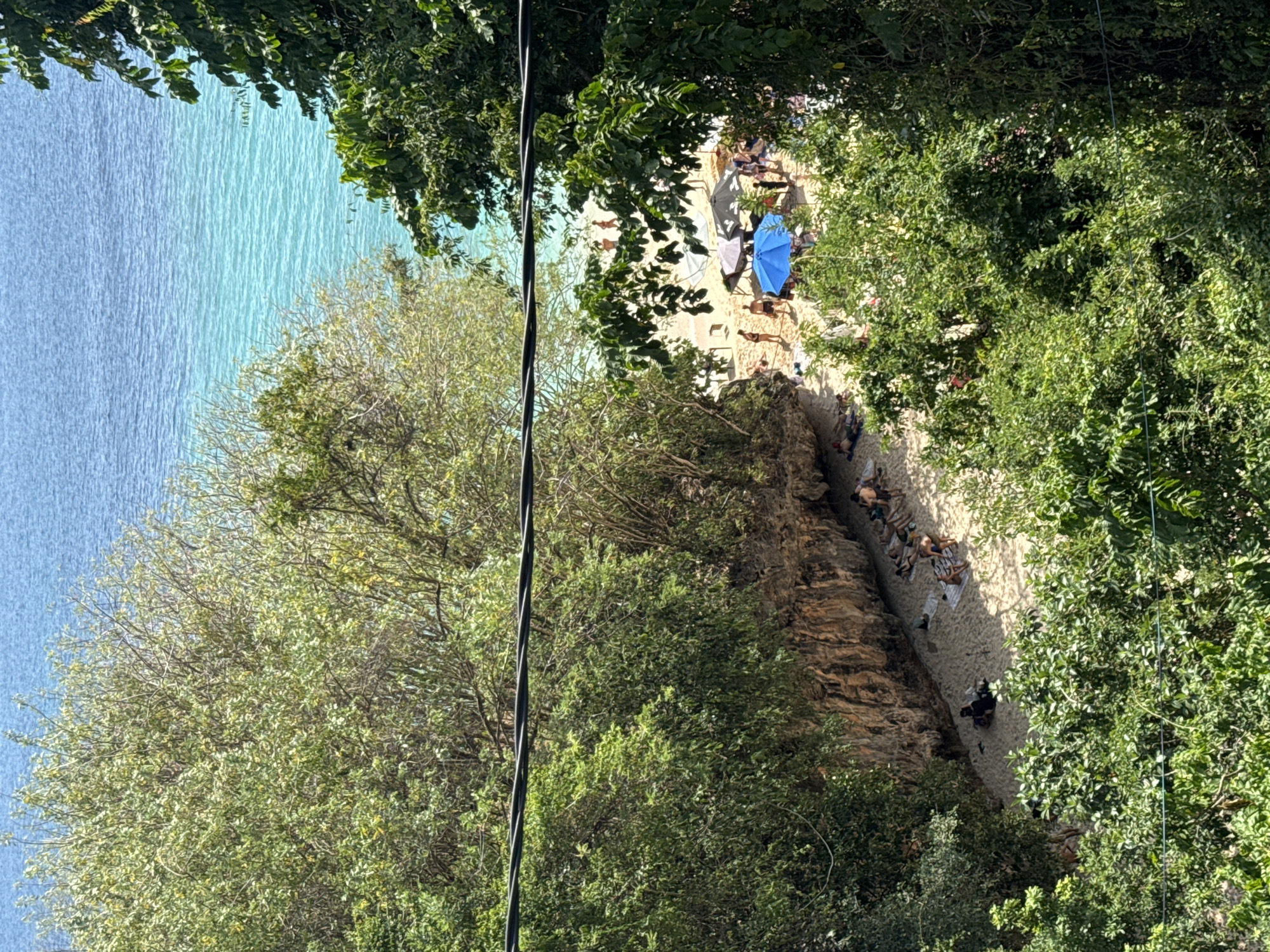 The image shows a tropical beach scene in Kuta Selatan, Indonesia, viewed from an elevated position through dense foliage. The beach is narrow, with light-colored sand, and bordered by a steep, rocky cliff face on the left and lush green trees on the right and foreground. The ocean in the background displays a gradient of blue and turquoise hues, with gentle waves.
Several people are visible on the beach, scattered in small groups. Some are sunbathing on towels, while others are standing or walking. There are several beach umbrellas providing shade, including a prominent bright blue one. The overall atmosphere suggests a relaxed day at the beach. A black cable is visible stretching horizontally across the upper portion of the image, obscuring some of the foliage and the sky.
