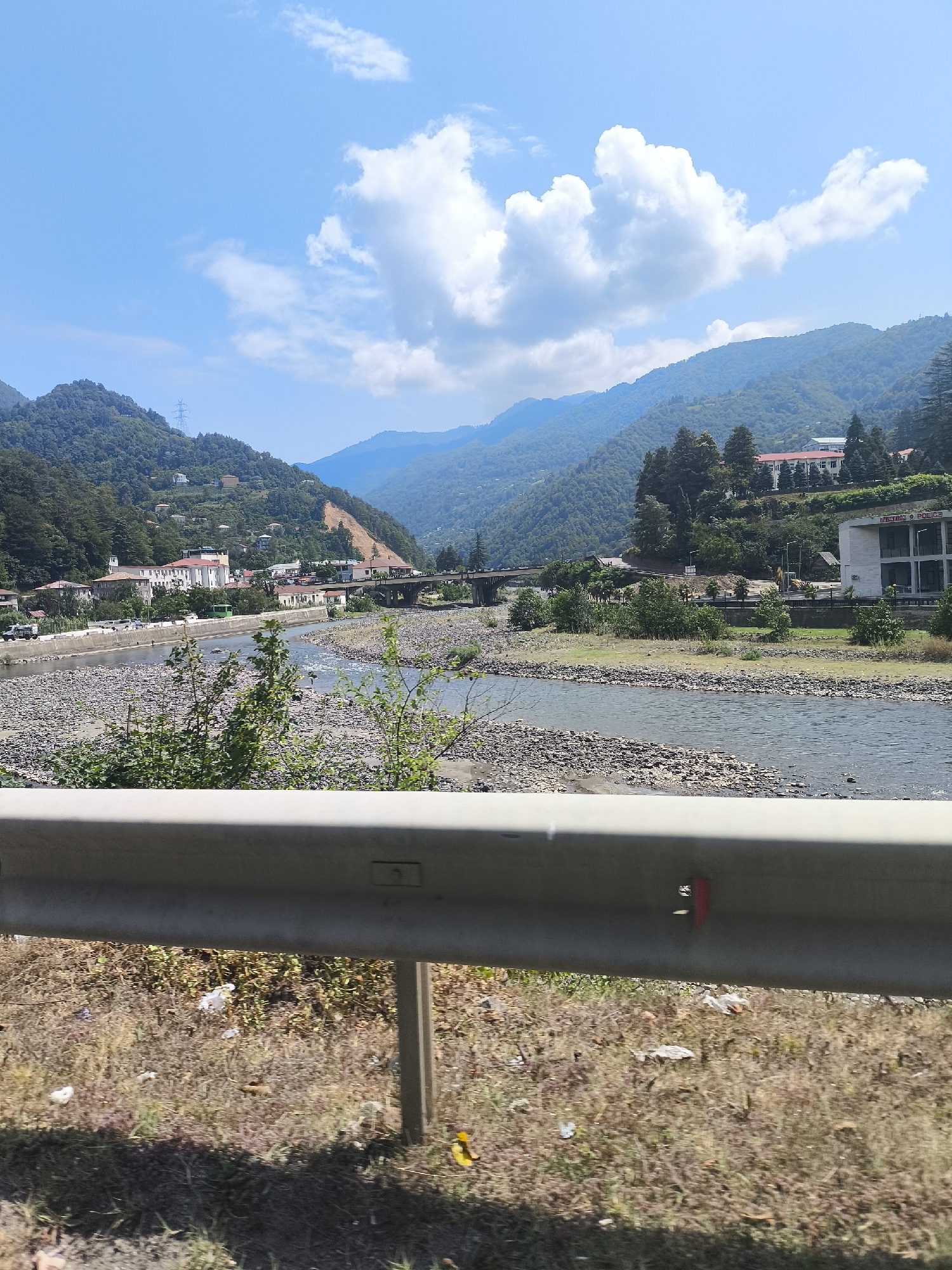 The image depicts a wide, shallow river flowing through a valley in Keda, Georgia. The riverbed is filled with numerous smooth, grey stones. On the left bank of the river, a series of buildings, some with white facades and red roofs, are nestled among green hills. A bridge crosses the river in the mid-ground, and more buildings and trees are visible on the right bank, with larger, darker green trees dominating the higher slopes. The sky above is a clear blue with scattered white clouds. In the foreground, a metal guardrail runs horizontally across the bottom of the frame, obscuring some of the dry, grassy embankment below.