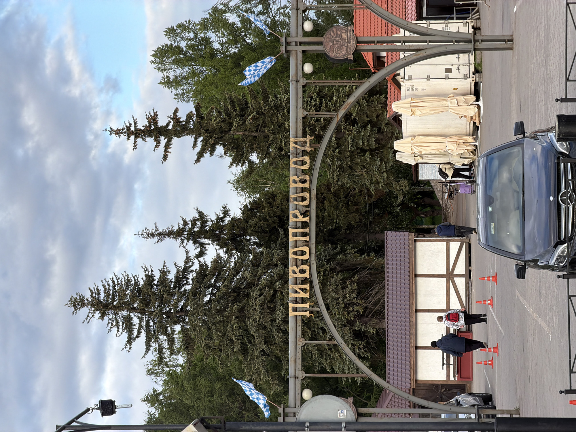 The image depicts an entrance gate in Saint Petersburg, Russia. The gate features the word "ПИВОПРОВОД" in gold Cyrillic letters. Bavarian flags are attached to the gate's upright poles. Behind the gate are tall evergreen trees and buildings with red tile roofs. A black Mercedes van is partially visible on the right side of the image. Orange cones are arranged in a line leading into the entrance. People are standing and walking near the entrance. In the background, shipping containers are stacked. The sky is overcast.