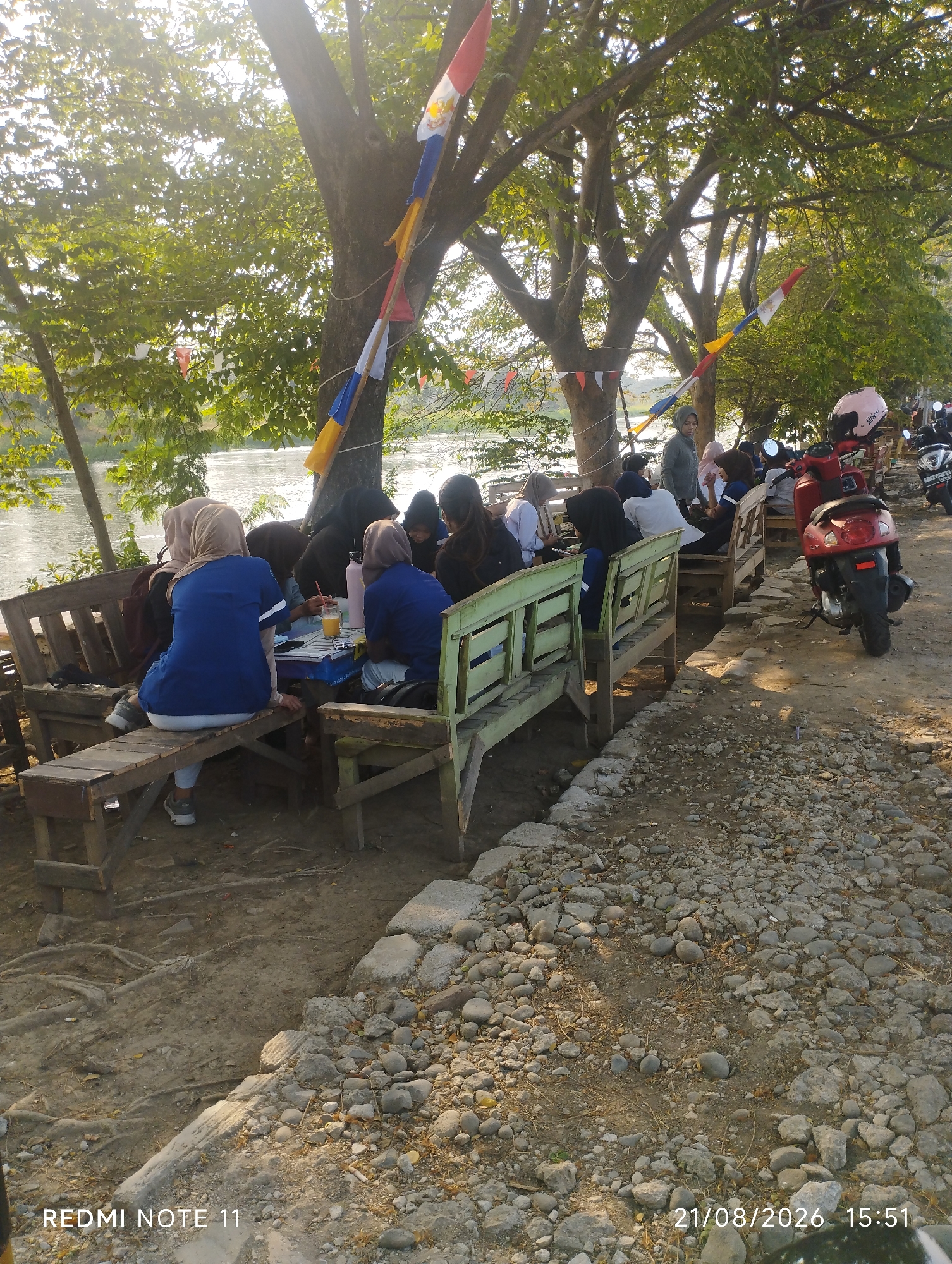 The image shows a group of people, primarily women wearing hijabs, gathered around wooden benches and tables near a body of water. They appear to be socializing or dining outdoors. Several motorcycles are parked in the background, suggesting a public gathering spot. The environment is natural, with trees and a river visible. Decorative flags are hung between trees, adding a festive touch. The scene captures a moment of leisure in what appears to be a riverside cafe or park, possibly in Karawang, Indonesia, indicated by the timestamp and the overall ambiance.