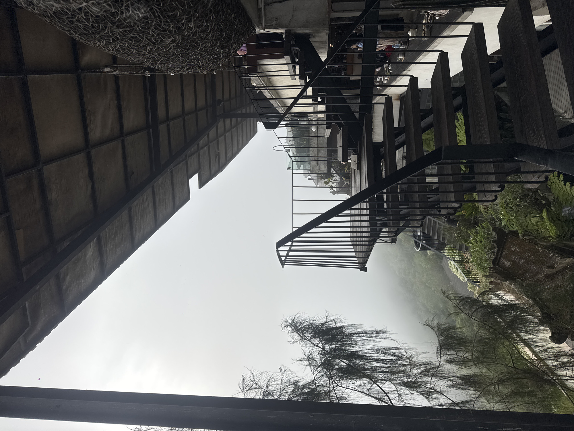 The image depicts an outdoor seating area with a view of a misty mountainous landscape, likely in Kintamani, Indonesia. A prominent feature is a dark metal spiral staircase with wooden steps, leading upwards and to the right. To the left, a large, dark vertical beam frames the scene, and a tree with wispy, drooping branches extends into the frame. Beyond the railing of the staircase, a lush green hillside is visible, shrouded in thick fog, with some wooden pathways and scattered vegetation. The upper portion of the image shows the underside of a roof structure, supported by dark metal beams, with a large woven decorative object hanging from the ceiling on the right. A few indistinct figures can be seen in the background near the stairs.