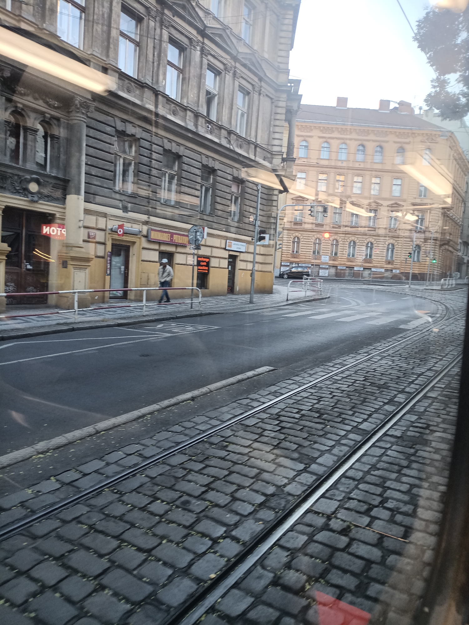 The image shows a street scene in Prague, Czechia, with tram tracks running along a cobblestone road. On the left, a building with ornate architecture and dark gray facade features multiple windows. A red neon sign with reversed text "ИОТЧ" is visible above a shop entrance. Further down, a person in a light-colored jacket and cap walks along the sidewalk, which is separated from the road by a metal railing. On the right, another building with a lighter, yellowish facade and arched windows is visible. The street is relatively empty, with only one black car visible in the distance. Traffic lights are present, with a red light illuminated. The perspective suggests the photo was taken from a moving tram.