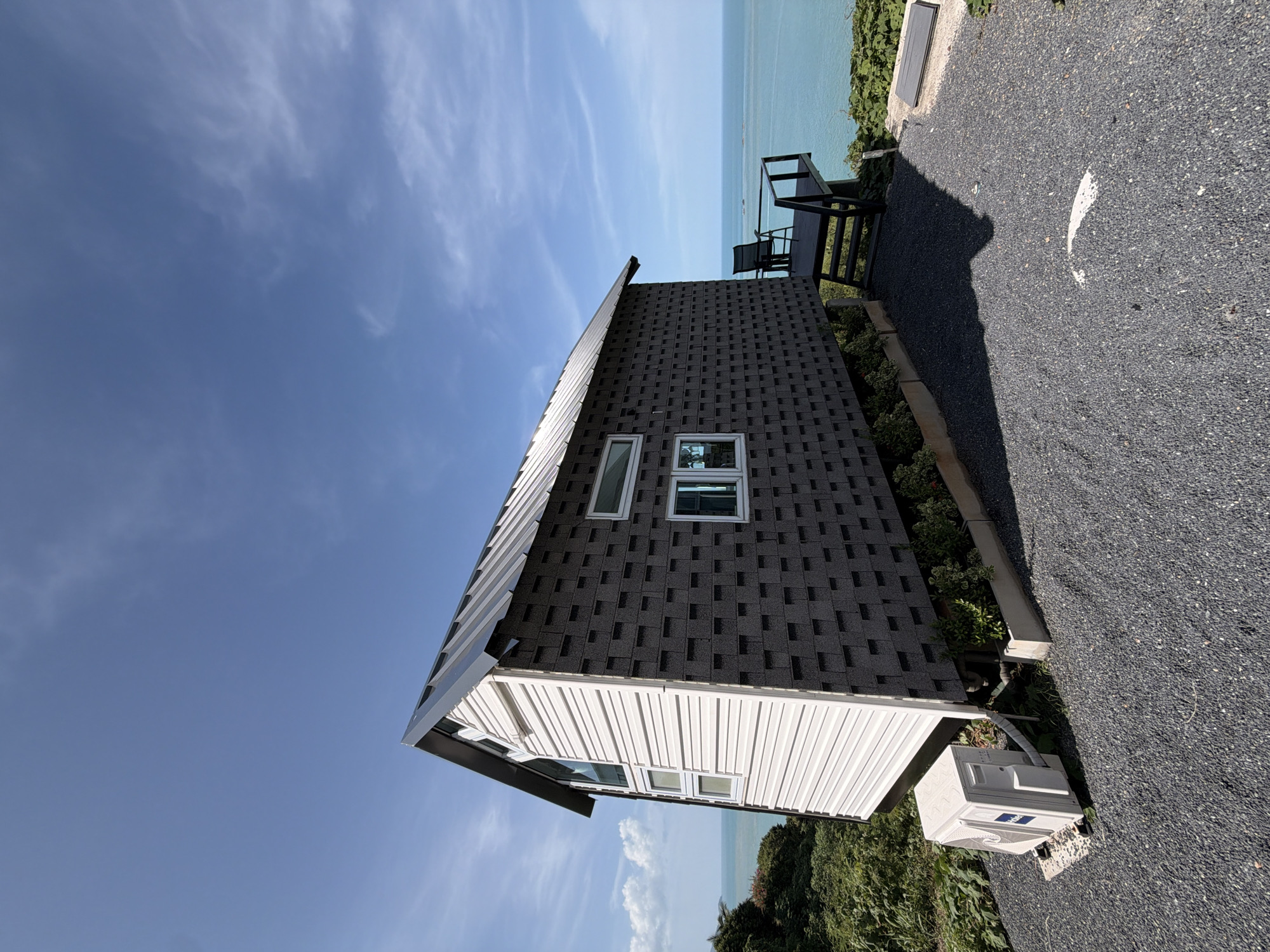 A low-angle, wide-angle shot captures a modern, rectangular, tiny house situated on a gravel driveway overlooking the ocean in Ko Samui District, Thailand. The house features a dark gray, shingled exterior on the main body, with a white, horizontally-paneled section on the left side. A dark metal roof slopes downwards, and several windows, some rectangular and some triangular, are visible. An outdoor air conditioning unit is positioned on the ground to the left of the house. To the right of the house, a small wooden deck with a black railing and a single chair extends towards the sea. The ocean, a vibrant turquoise, stretches to the horizon under a bright blue sky with scattered white clouds. Lush green vegetation surrounds the house, and the ground is covered in dark gray gravel.