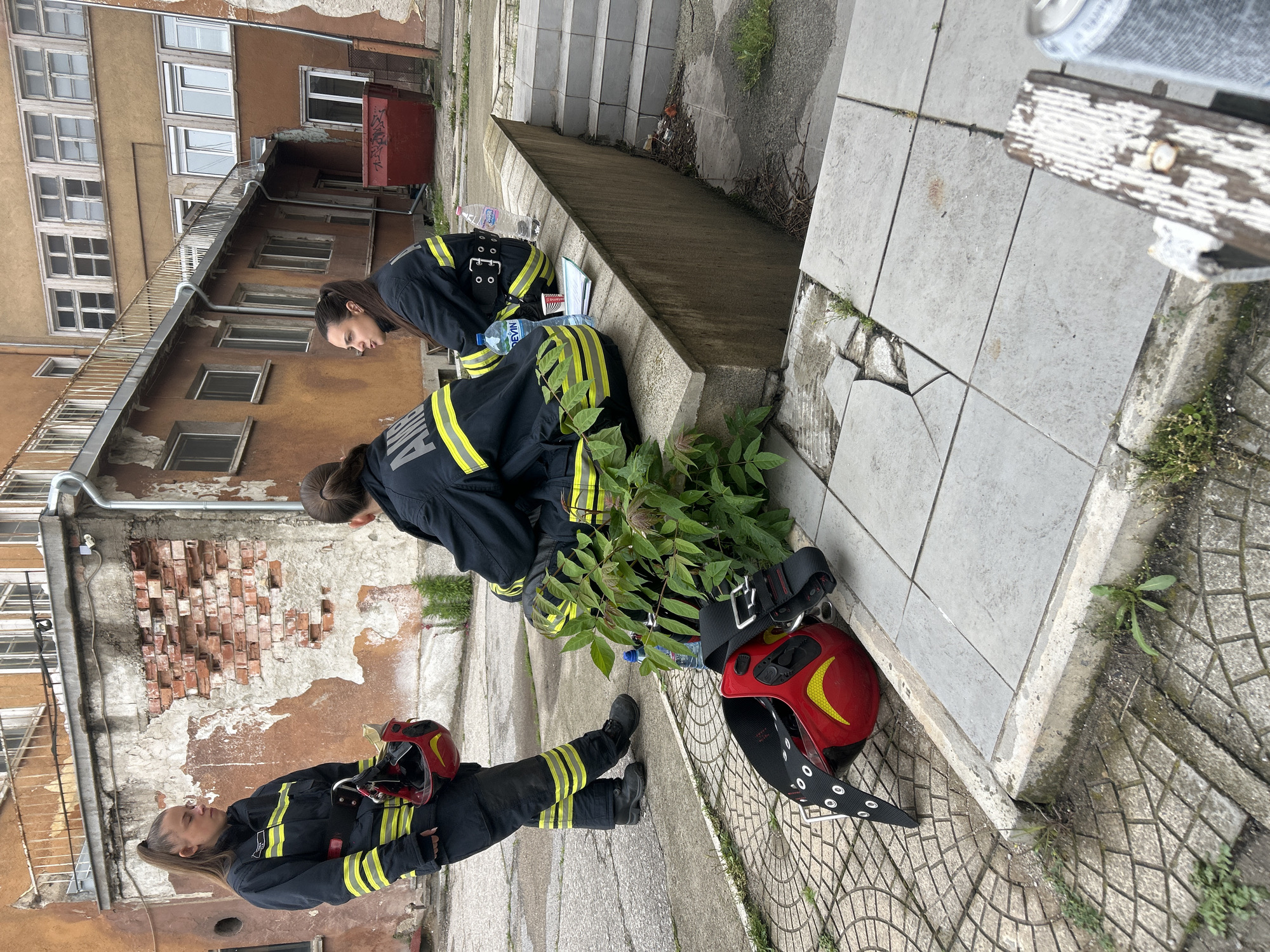 This image depicts three individuals, appearing to be female firefighters, taking a break in an urban setting in Sofia, Bulgaria. Two are seated on a concrete ledge or step, while one stands in the foreground, holding her red firefighter helmet.
All three are dressed in dark navy or black firefighter uniforms, distinguished by prominent yellow and silver reflective stripes across the shoulders, arms, and legs. Two red helmets, each adorned with a yellow crescent emblem, are visible on the ground beside the seated individuals. Water bottles, at least one with the brand "DEVIN" visible, are placed near the seated figures. The uniform of the standing person clearly displays "SOFIA" on the back, while the seated person closest to the camera has text resembling "ПОЖАРНА" (Bulgarian for "Fire Department") on her uniform.
The scene appears to be an outdoor alleyway or courtyard between old, multi-story buildings. These buildings exhibit signs of age with crumbling facades, peeling paint, exposed brickwork, and numerous windows. A metal railing is visible along a lower roofline. The ground is paved with a mix of concrete slabs and patterned tiles, with sections of cracked pavement and weeds growing in crevices. Steps lead up in the background. The lighting suggests it is daytime, possibly under an overcast sky, creating diffused light without harsh shadows. The individuals appear to be resting, perhaps after an activity or on standby.