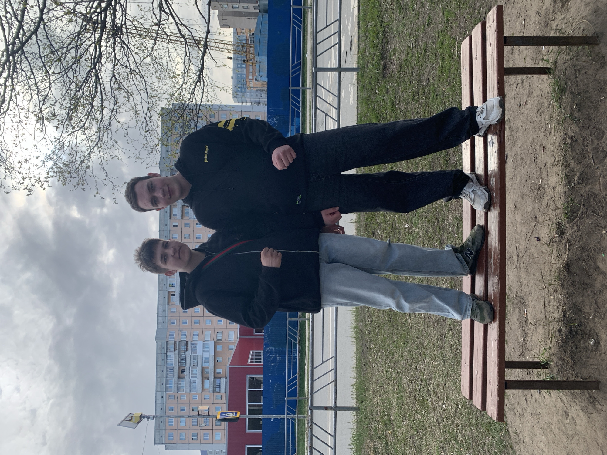 Two young men stand in front of a residential building in Severodvinsk, Russia, with their feet on a brown park bench. The man above wears a black jacket with yellow stripes on the sleeves, black pants, and white sneakers. The man below wears a black jacket, light blue jeans, and green sneakers. Behind them is a sidewalk with a blue metal fence and a building with red and white sections. A bare tree is visible in the upper left corner, and the sky is overcast.