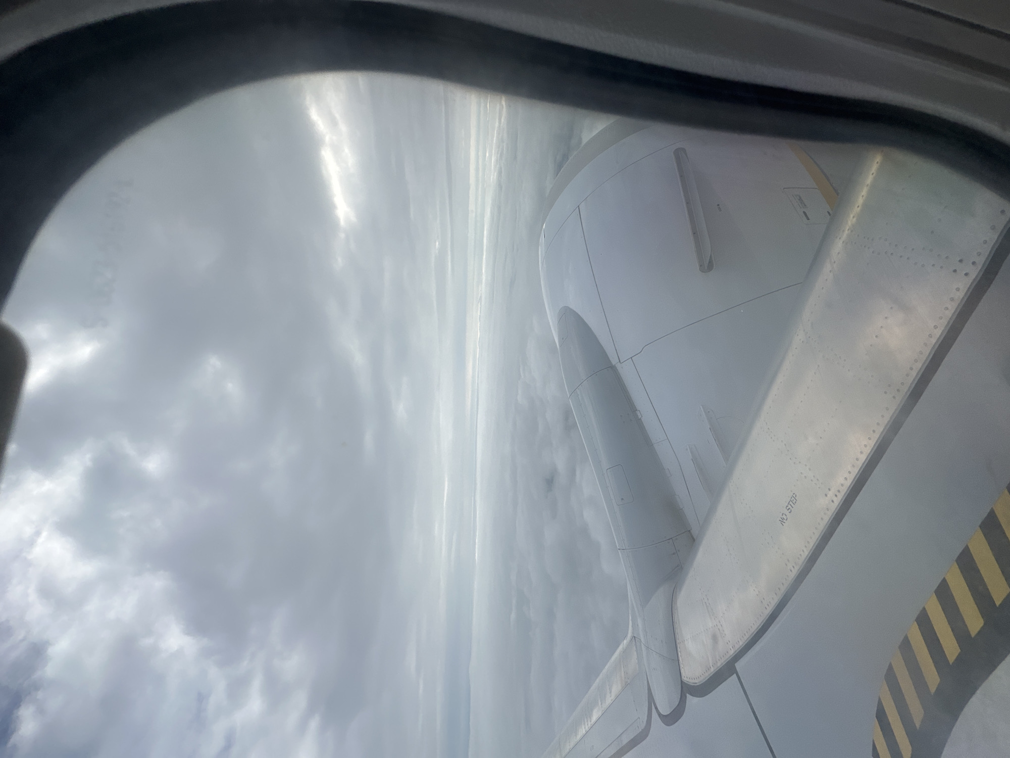 An aerial view from an airplane window shows a vast expanse of clouds below and an overcast sky. The wing of the aircraft, made of metal panels with visible rivets, extends into the frame, featuring a black and yellow striped anti-slip marking. Part of an aircraft engine nacelle is also visible. The scene is dominated by shades of gray and white, with diffused sunlight breaking through the clouds. No people are visible. There is no indication of Krivina, Bulgaria.