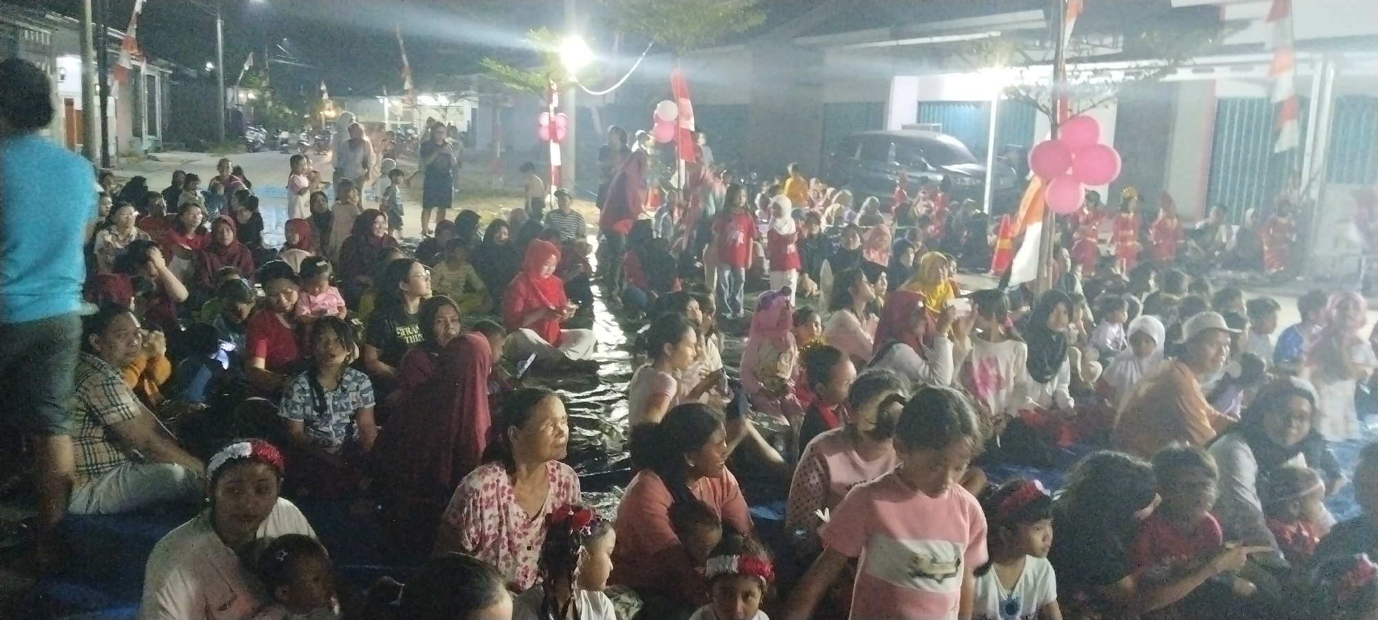 This is a wide-angle, medium-shot, outdoor photograph taken at night, possibly during a community event or celebration in Taban, Indonesia. A large crowd of people, predominantly women and children, are seated on the ground, facing towards the right of the frame, where some performers in traditional red and gold attire appear to be on a stage or elevated area. The lighting is dim and artificial, with some bright spotlights illuminating the performers and casting long shadows.
Flags, likely Indonesian flags, are visible on poles, adorned with red and white stripes. Several pink balloons are tied to a pole in the mid-ground, adding a festive touch. In the background, there are buildings, some with lights on, and a vehicle is parked. The ground appears to be wet or reflective, possibly due to recent rain or a water feature.
The individuals in the crowd are diverse in age and appearance, dressed in casual clothing, with many women wearing headscarves. The atmosphere seems to be one of communal gathering and enjoyment of a performance. The overall impression is of a lively local event in a residential area at night.