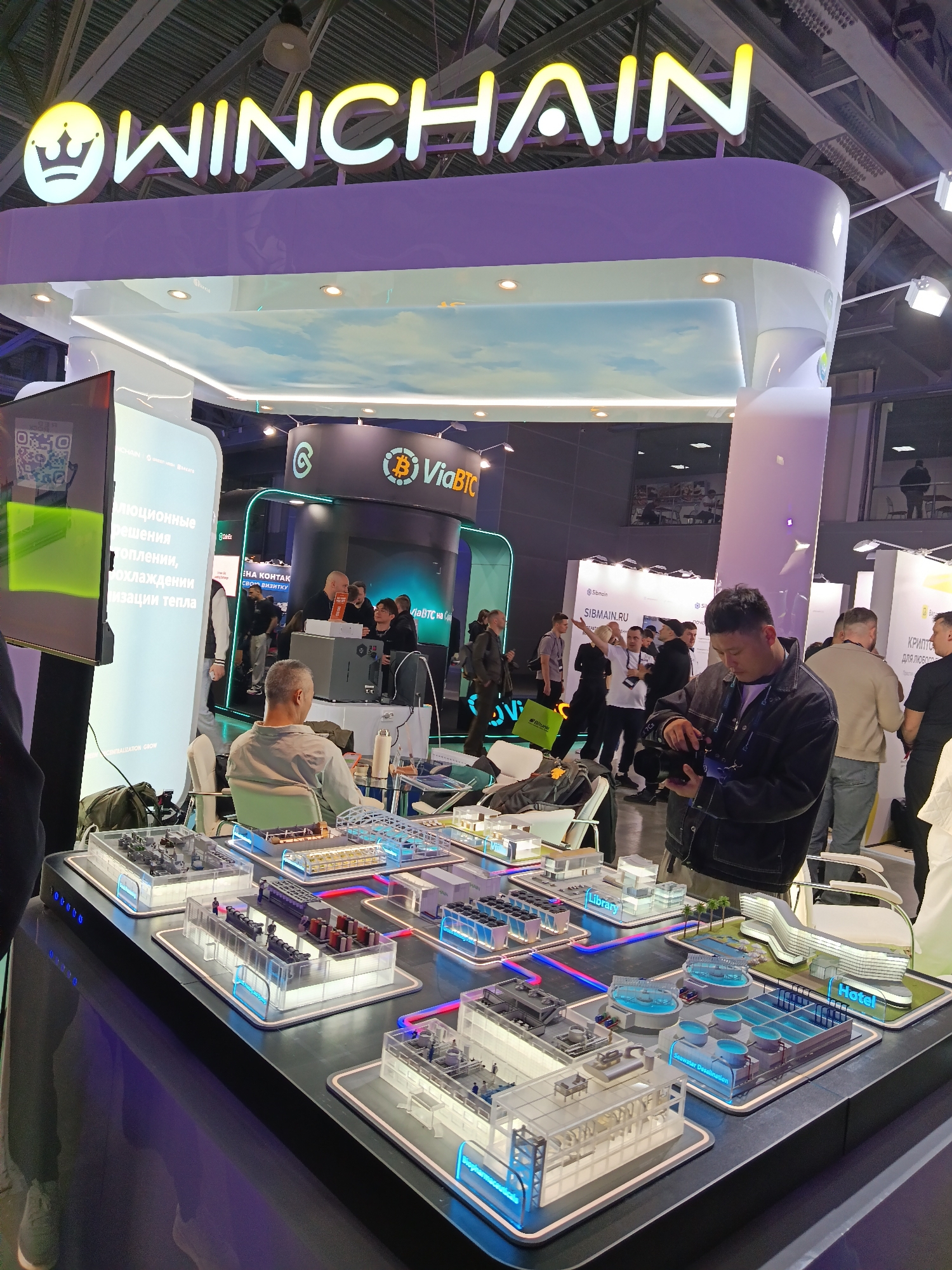 This image was taken at a trade show in Khimki, Russia. The foreground features a modern exhibit with a black tabletop displaying multiple miniature models of industrial and residential infrastructure, including a hotel, library, and seawater desalination plant, each outlined with vibrant LED lights. A man in a dark jacket is examining a camera or phone, possibly taking a photo of the exhibit. Behind him, various attendees are walking and gathering near other booths, including one with the "ViaBTC" logo. A large sign overhead reads "WINCHAIN" in yellow and white illuminated letters, with a crown emblem above the "W". The environment is well-lit and bustling with activity.